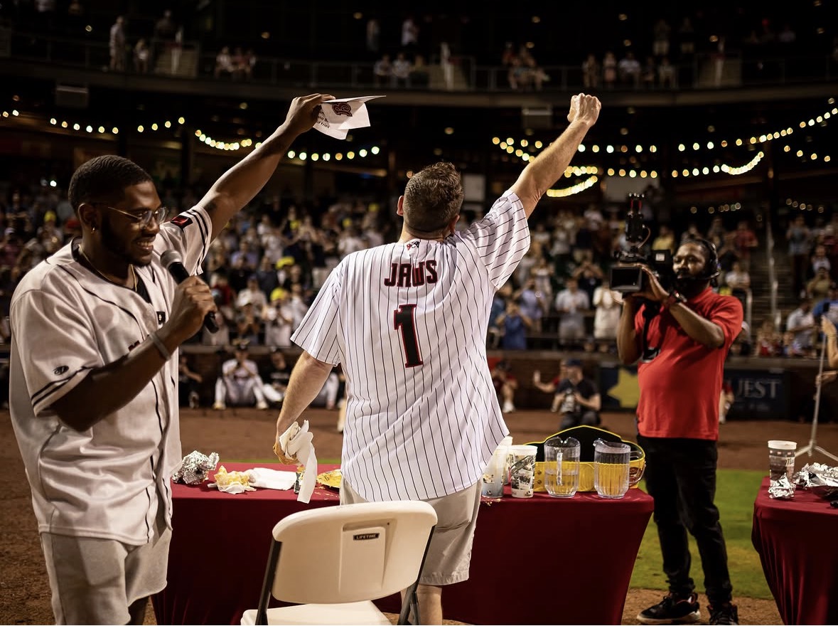 Person in a baseball jersey with number 1 raises arm, others around, in a stadium with audience, celebrating.