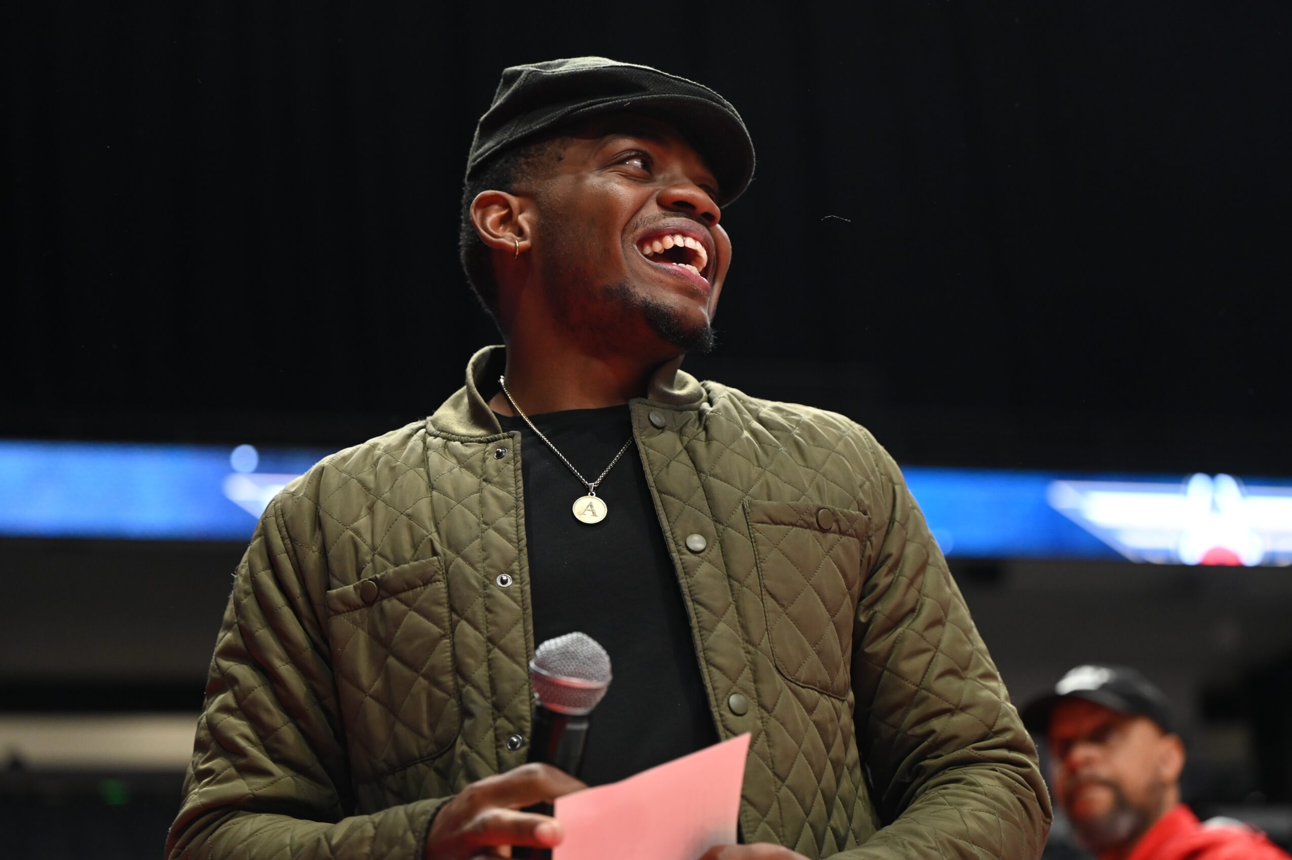Man smiling, holding a microphone and paper, wearing a cap and quilted jacket, in an indoor setting.