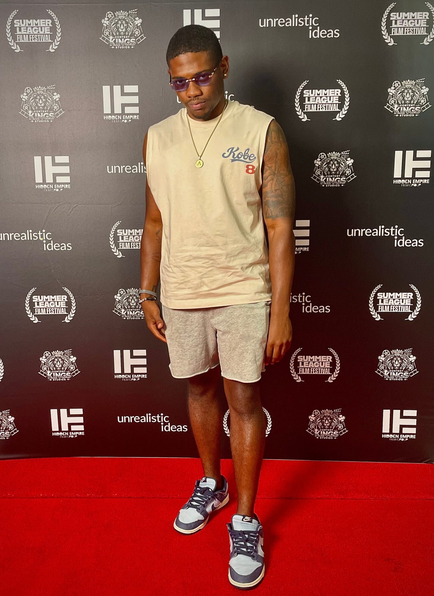 Young man wearing sunglasses, sleeveless shirt, shorts, and sneakers standing against a black backdrop with logos.