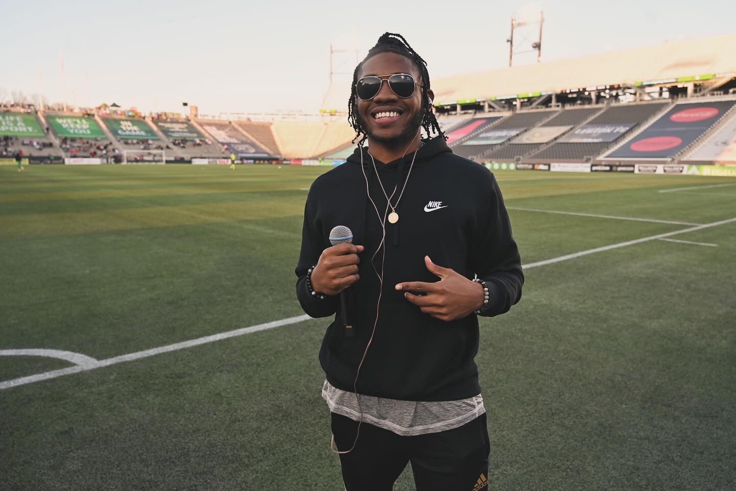 Person smiling, holding a microphone, standing on a sports field with empty stadium seats in background.