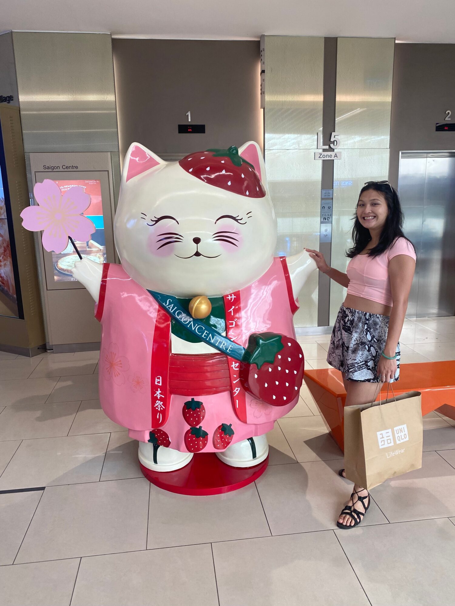Woman standing next to a large, pink, cat-shaped statue with a blue bow, in an indoor setting.
