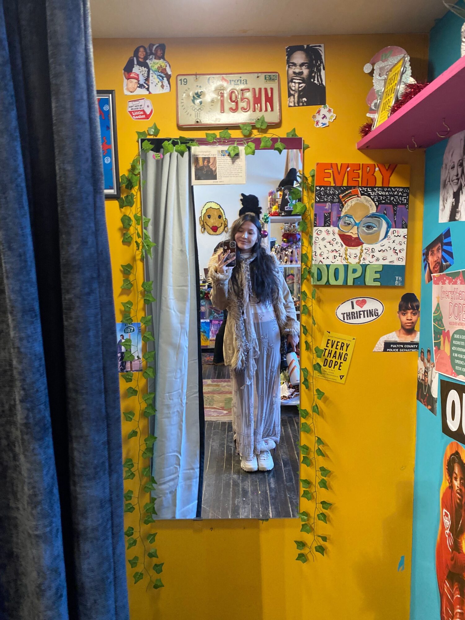 Person standing in front of a colorful wall with various posters and decorations, wearing a long dress and holding a bag.