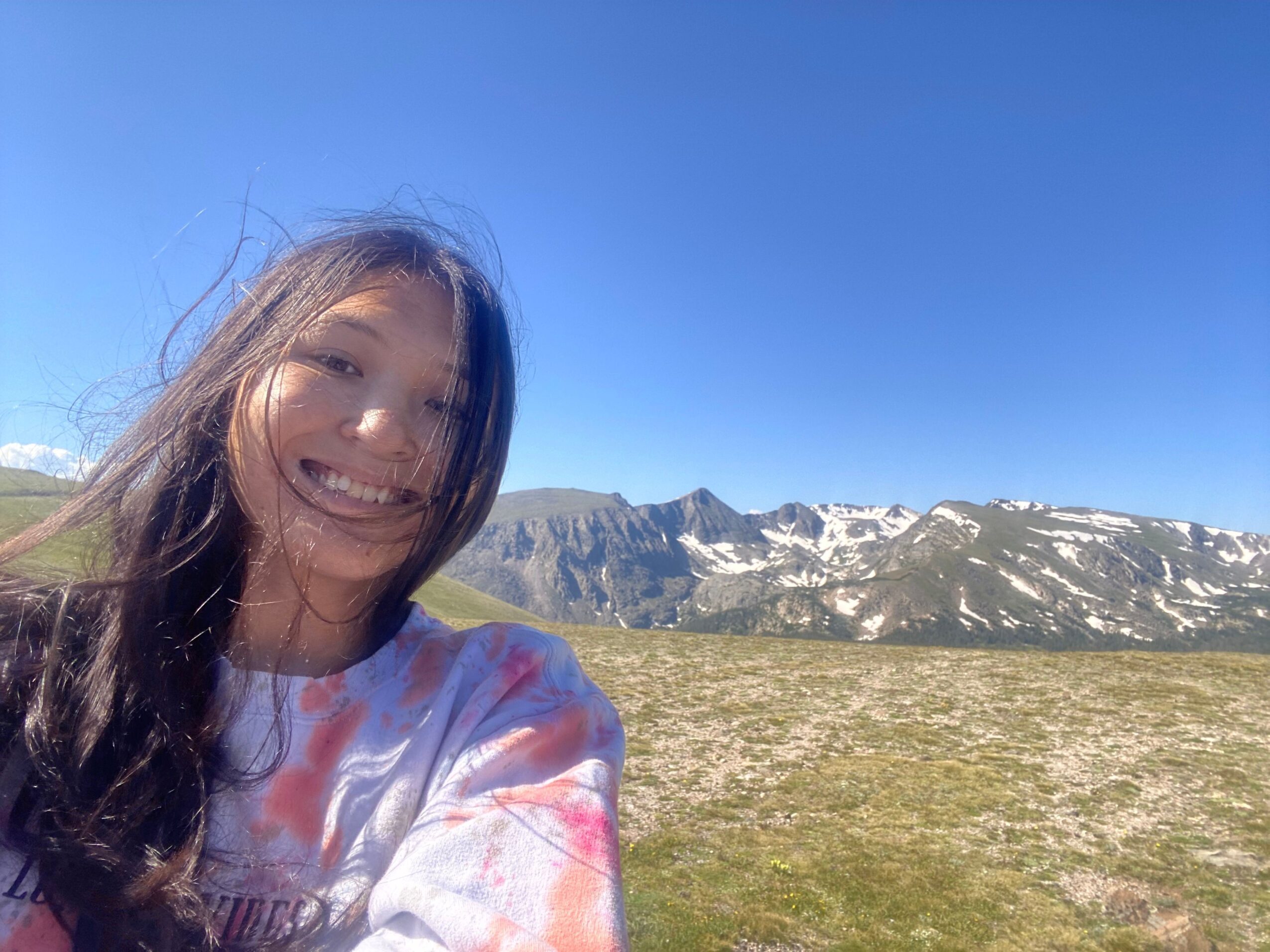 Girl with long hair and glasses smiling outdoors with mountains and blue sky in background.