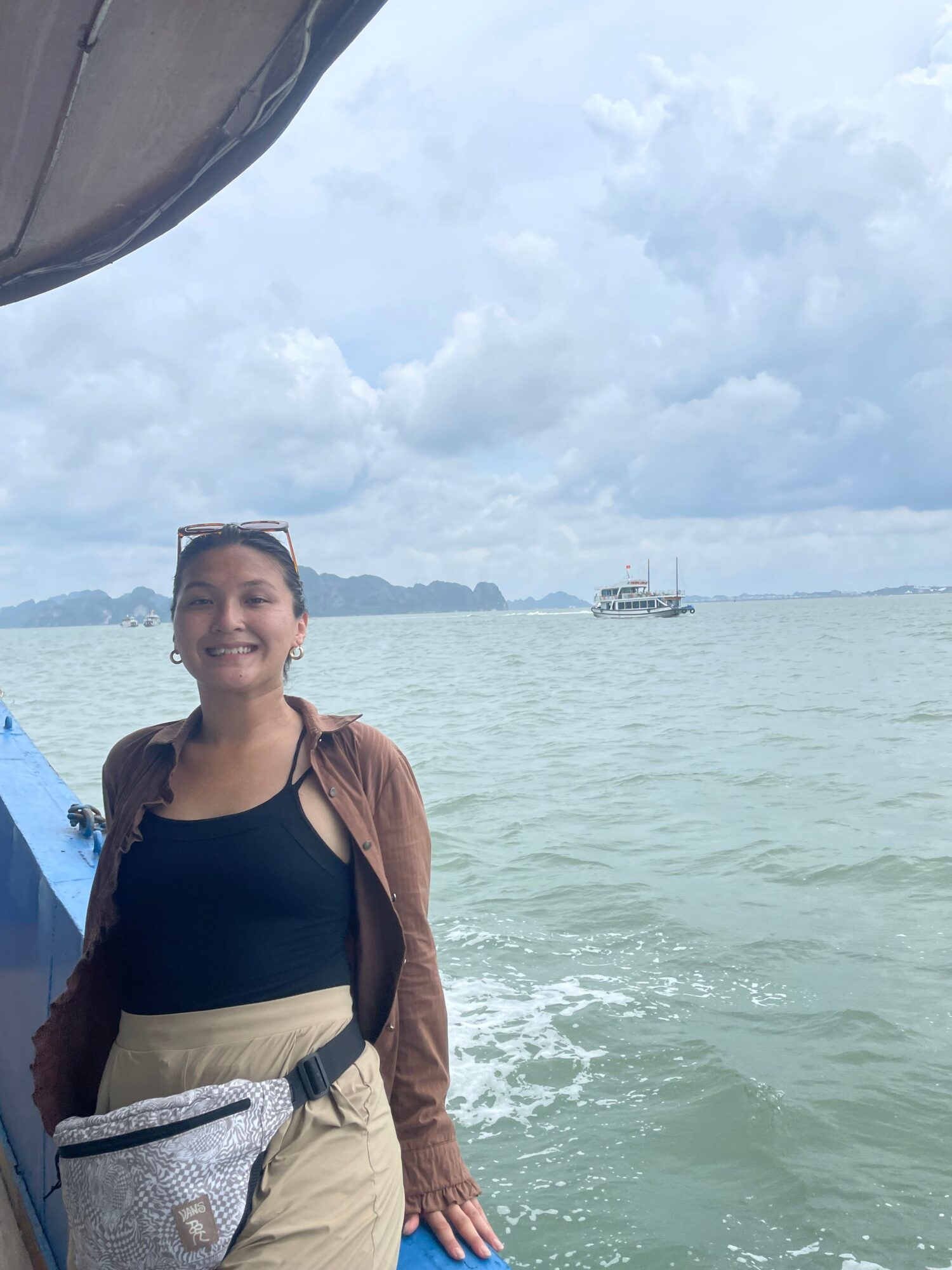 Woman standing near water with a boat in the background, cloudy sky, wearing a brown jacket and black top.