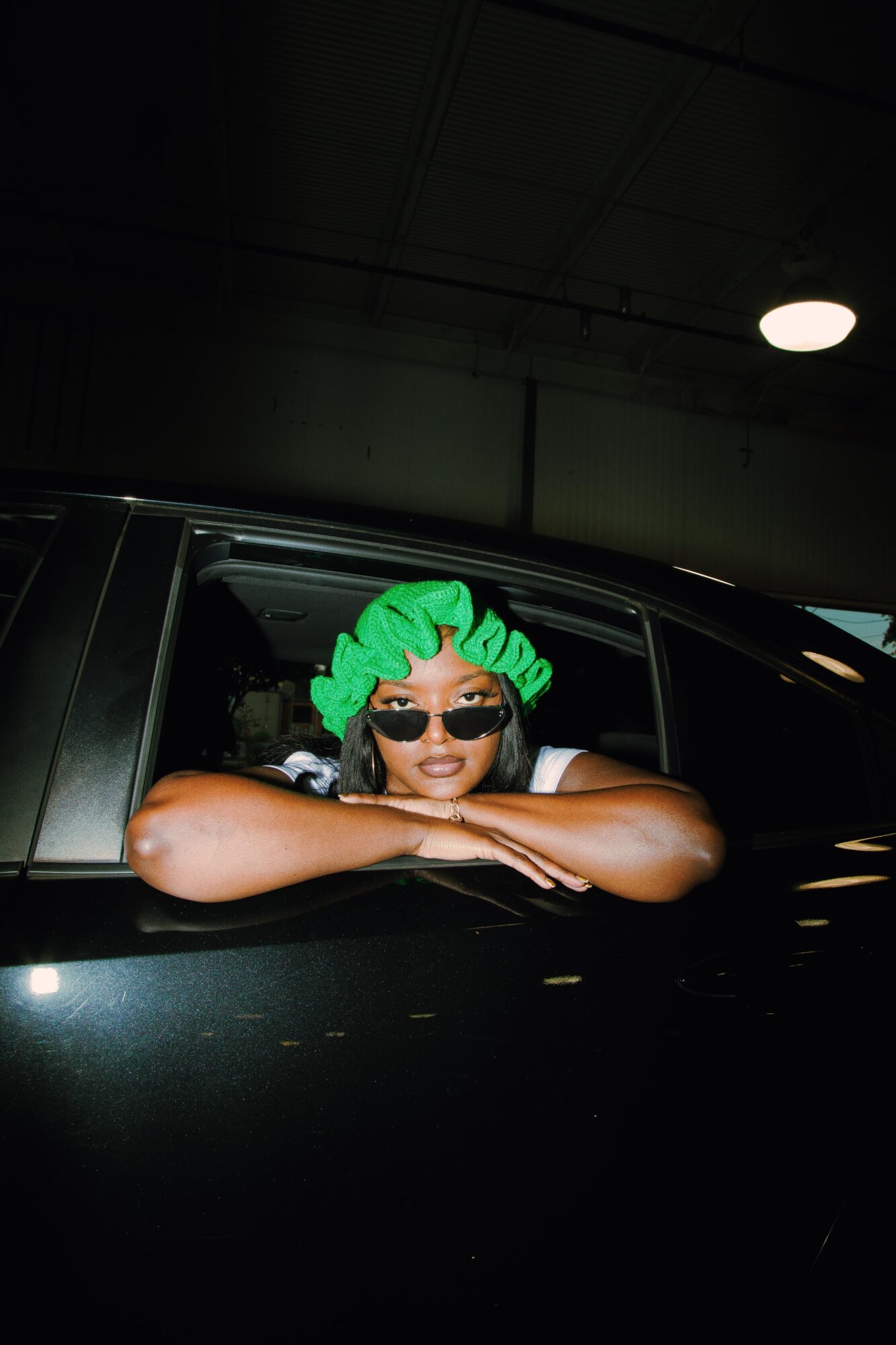 Person with green hair and sunglasses leaning on a car window in an indoor parking garage.