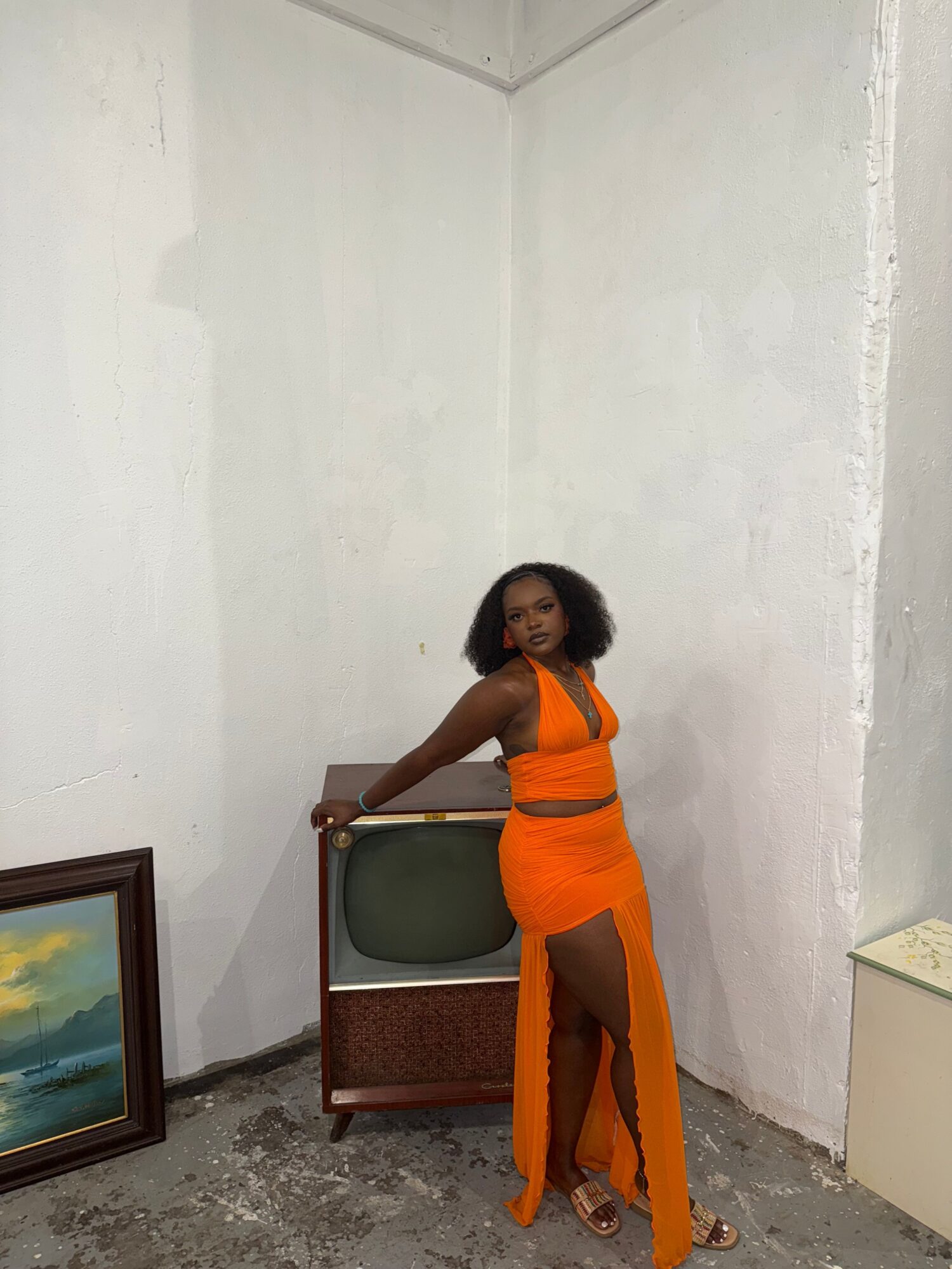 Woman in orange dress leaning on a chair in a room with white walls and a framed painting.