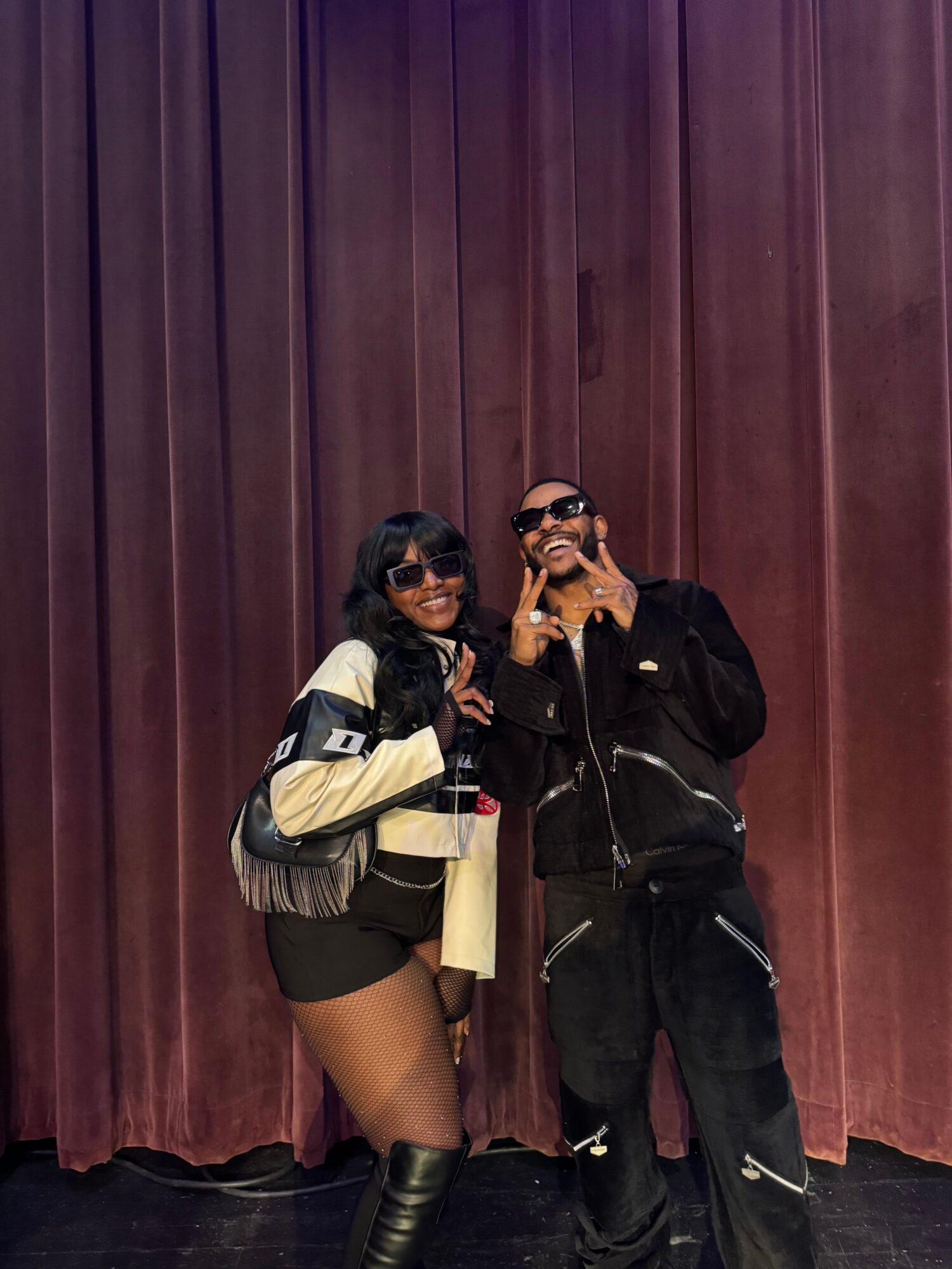 Two people taking a selfie in front of a maroon curtain, one sitting and the other standing, both wearing dark clothing and masks.