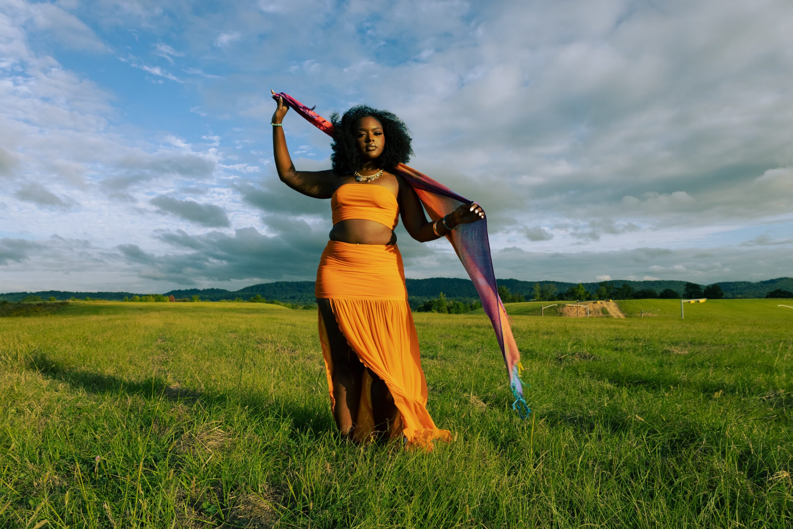 Woman standing in a grassy field holding a colorful scarf, with a cloudy sky and distant landscape in the background.