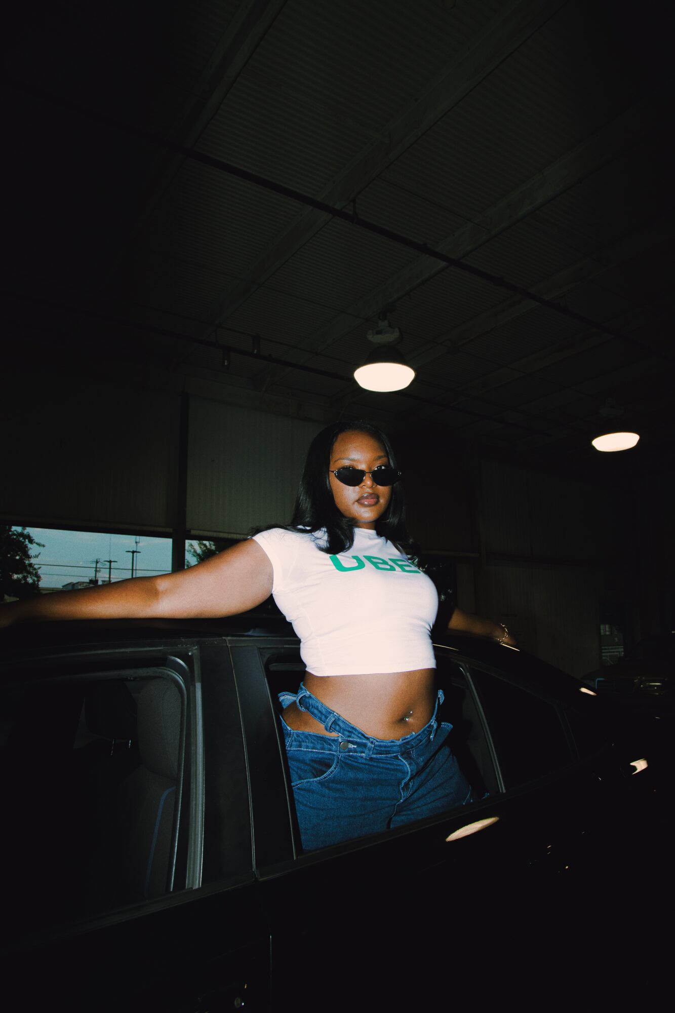 Woman with sunglasses leaning on a car in a dark parking garage, wearing a white crop top and jeans.