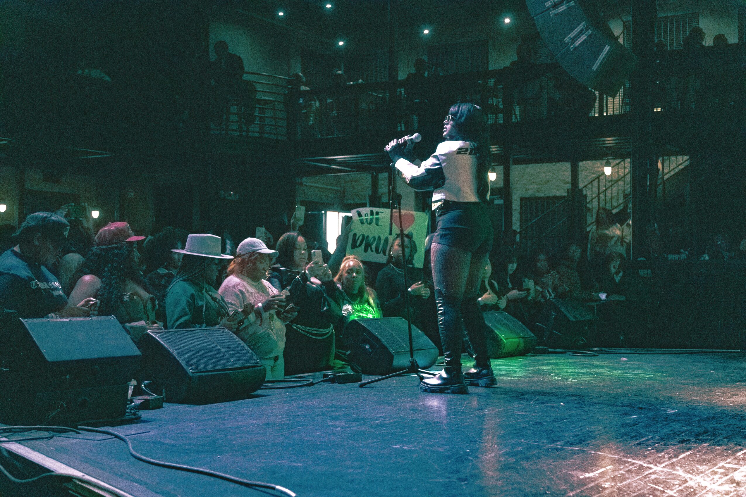 Performer on stage with audience seated in front, stage lights shining, in a dark indoor venue.