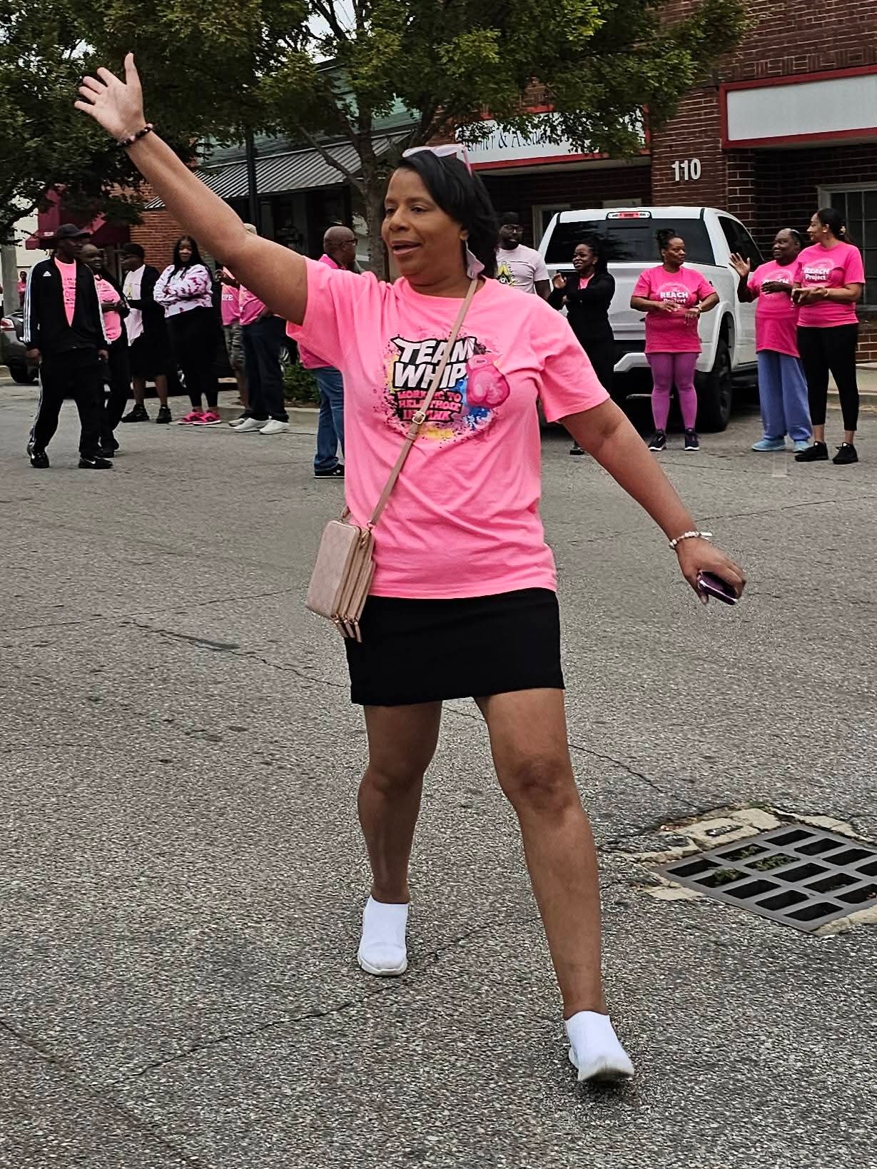 Woman in pink T-shirt and black skirt waving on street with group of people in pink shirts in background.