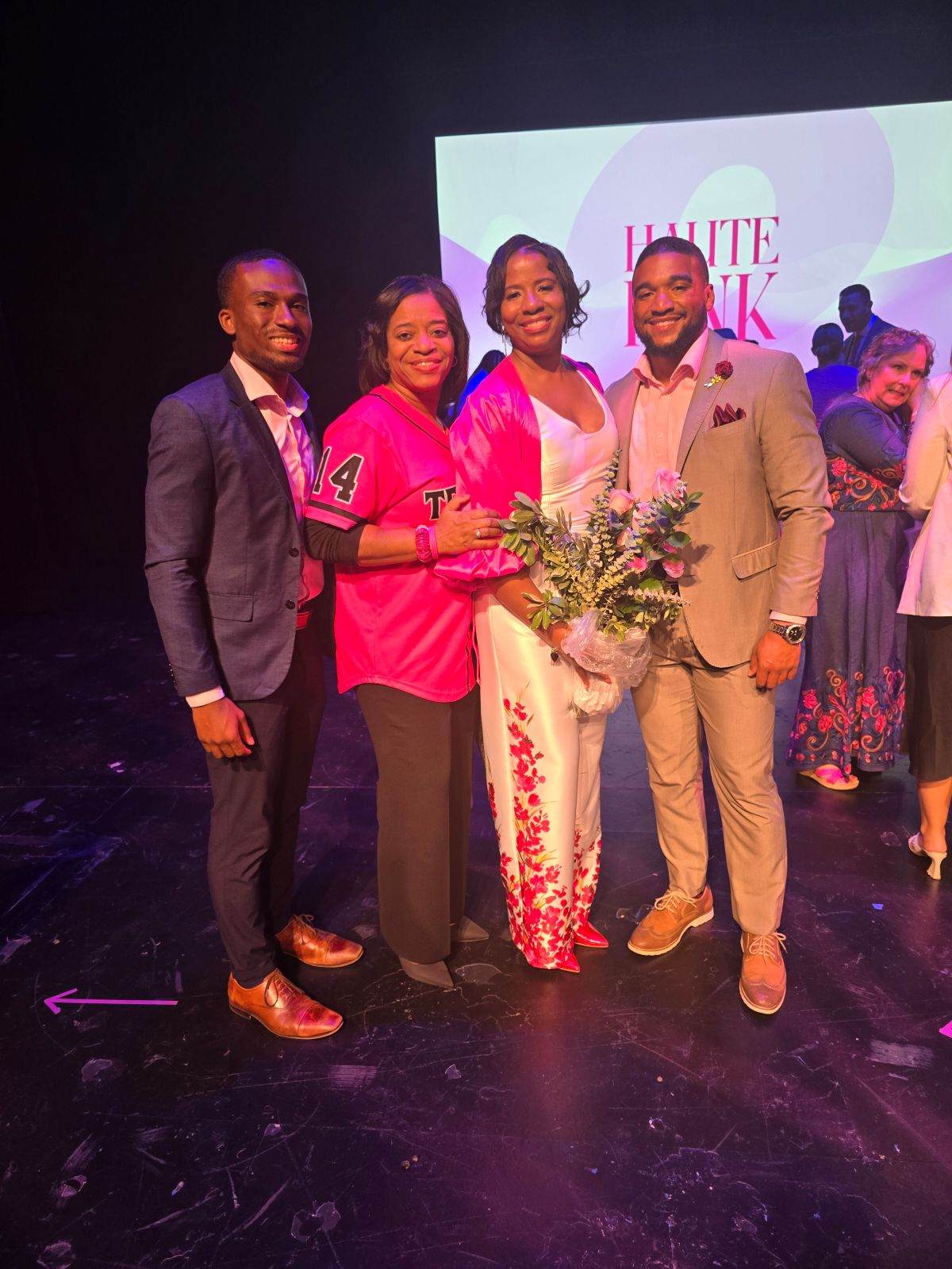 Four people stand on stage, smiling, with a screen behind them displaying text. One person holds a bouquet of flowers.