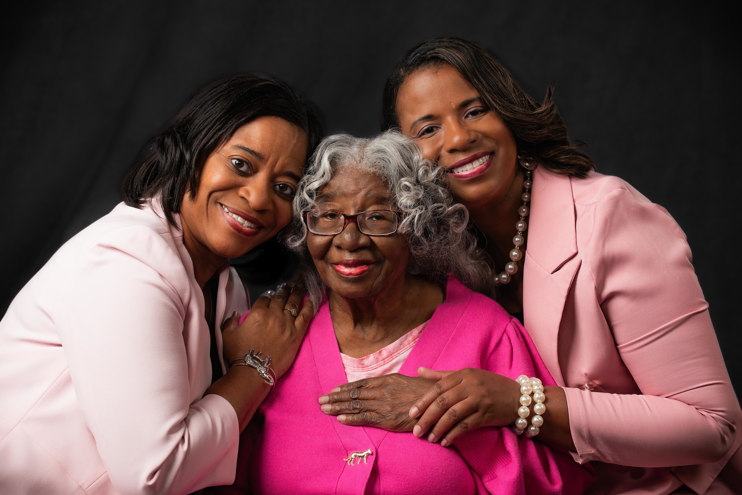 Three women, one elderly and two younger, smiling and embracing each other, against a dark background.