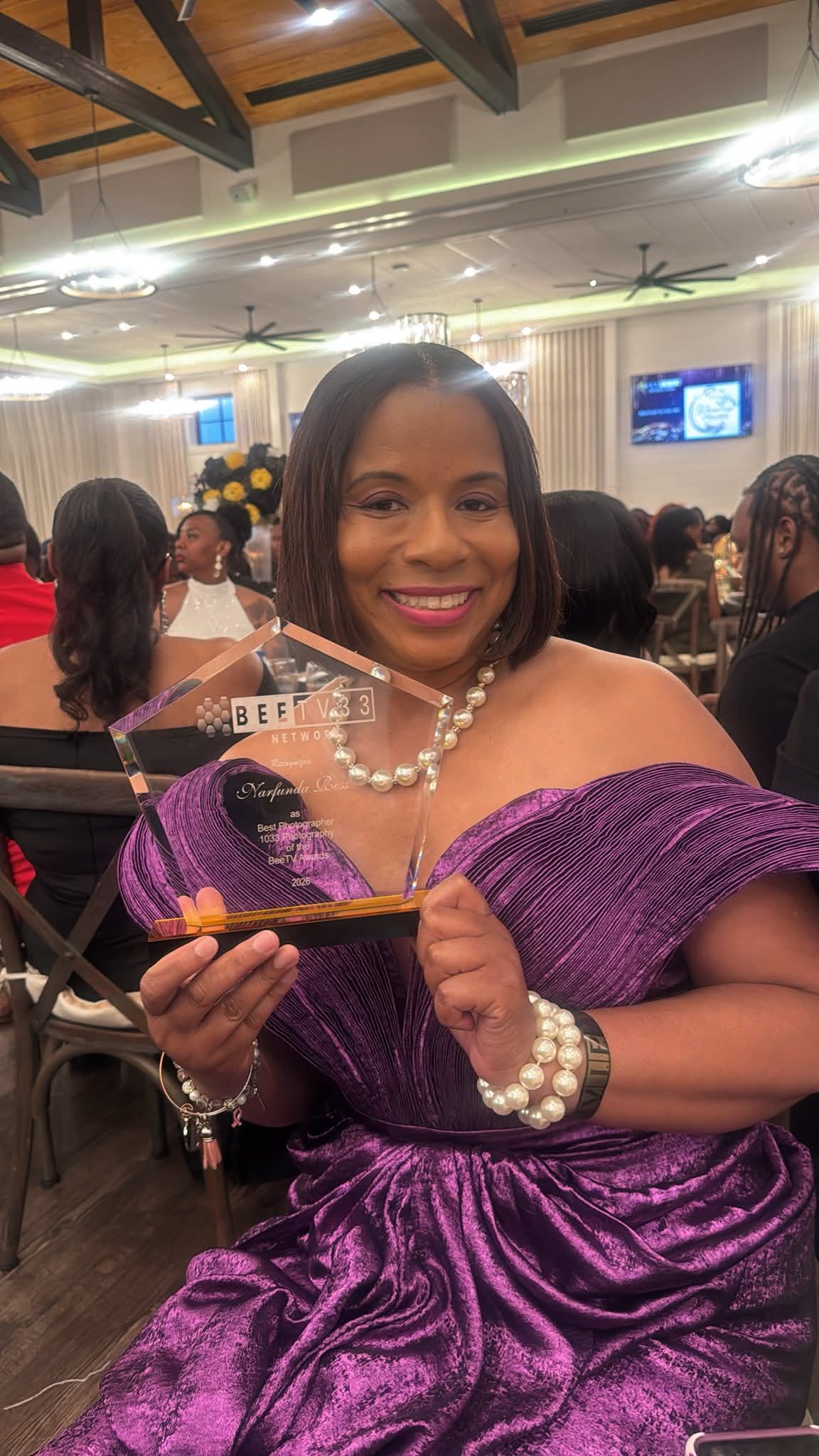 Young woman in purple dress with pearl necklace and bracelet, smiling at a formal event, holding a small object.