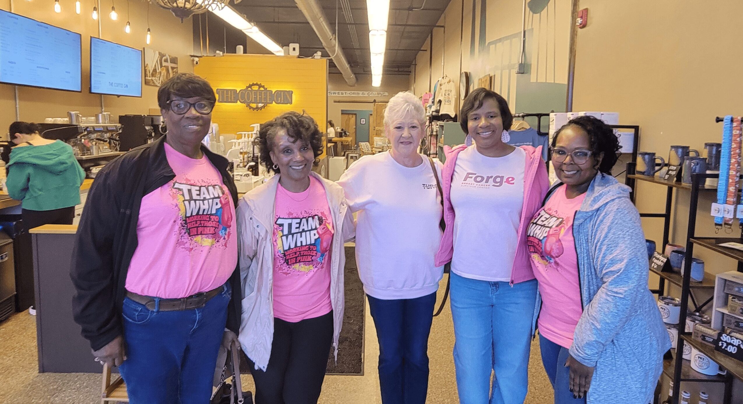 Group of five women standing together indoors, some wearing pink shirts with text, smiling at the camera.