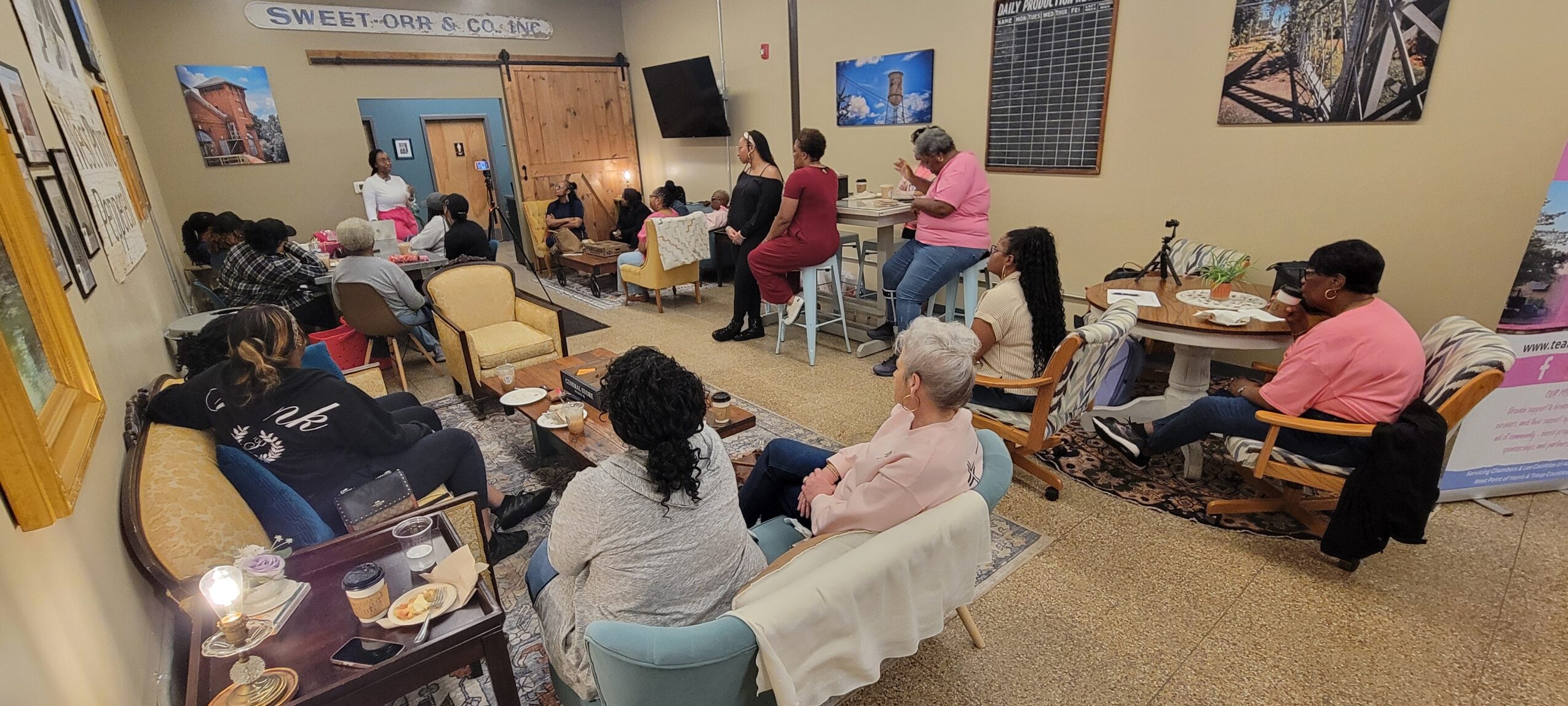 Group of people gathered in a room, some sitting on chairs and couches, others standing, engaging in a discussion or activity.