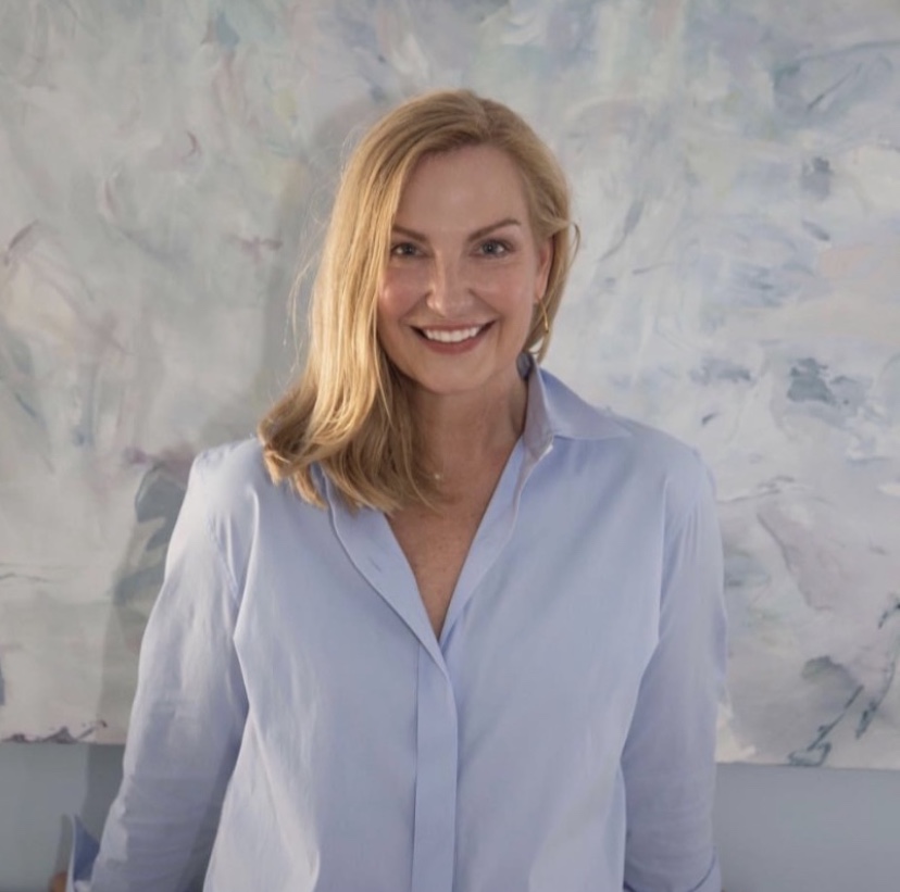 Woman with shoulder-length blonde hair smiling, wearing a light blue shirt, standing in front of an abstract background.