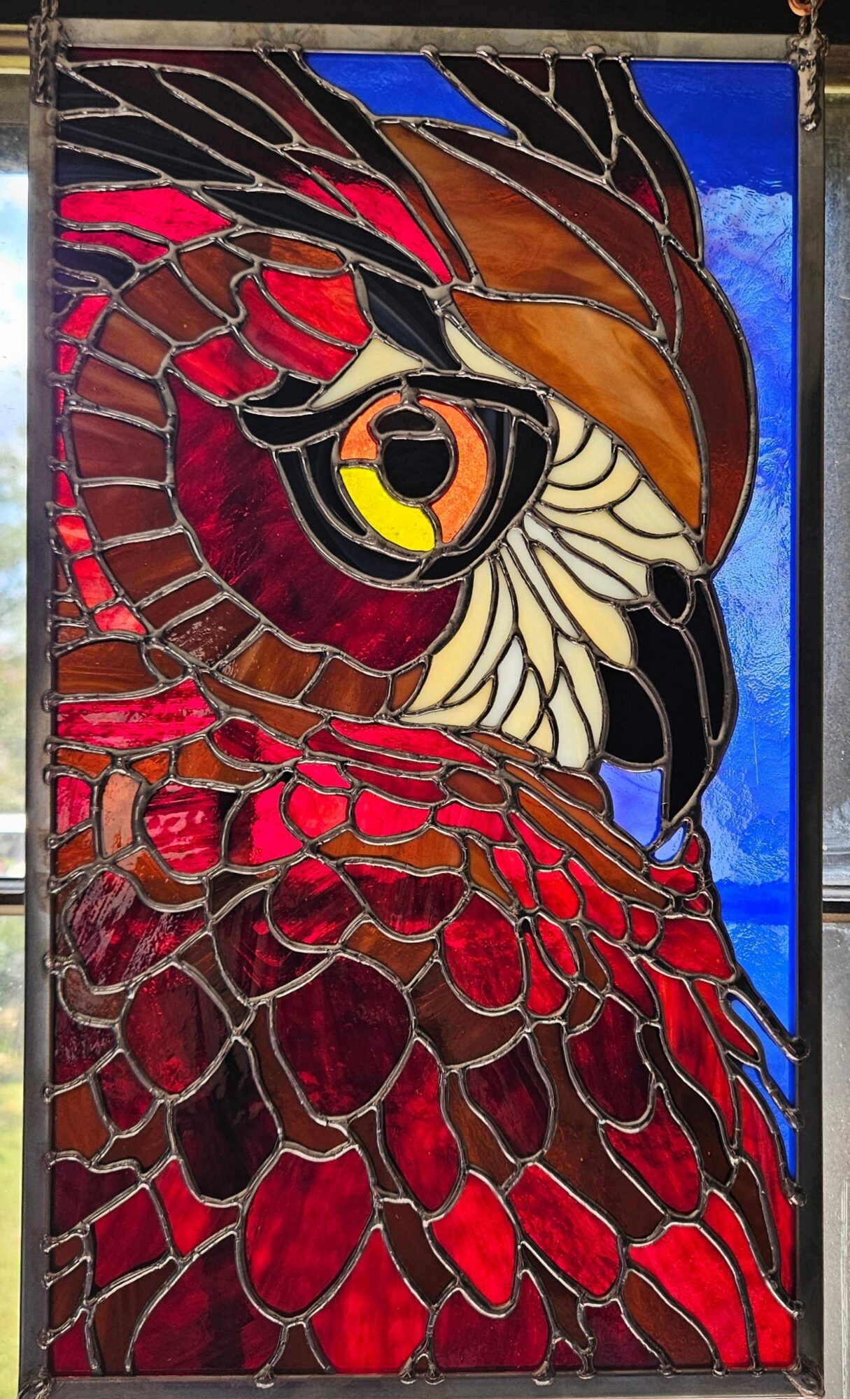 Colorful stained glass window depicting a bird with red, brown, white, and black feathers, and a yellow eye, against a blue background.
