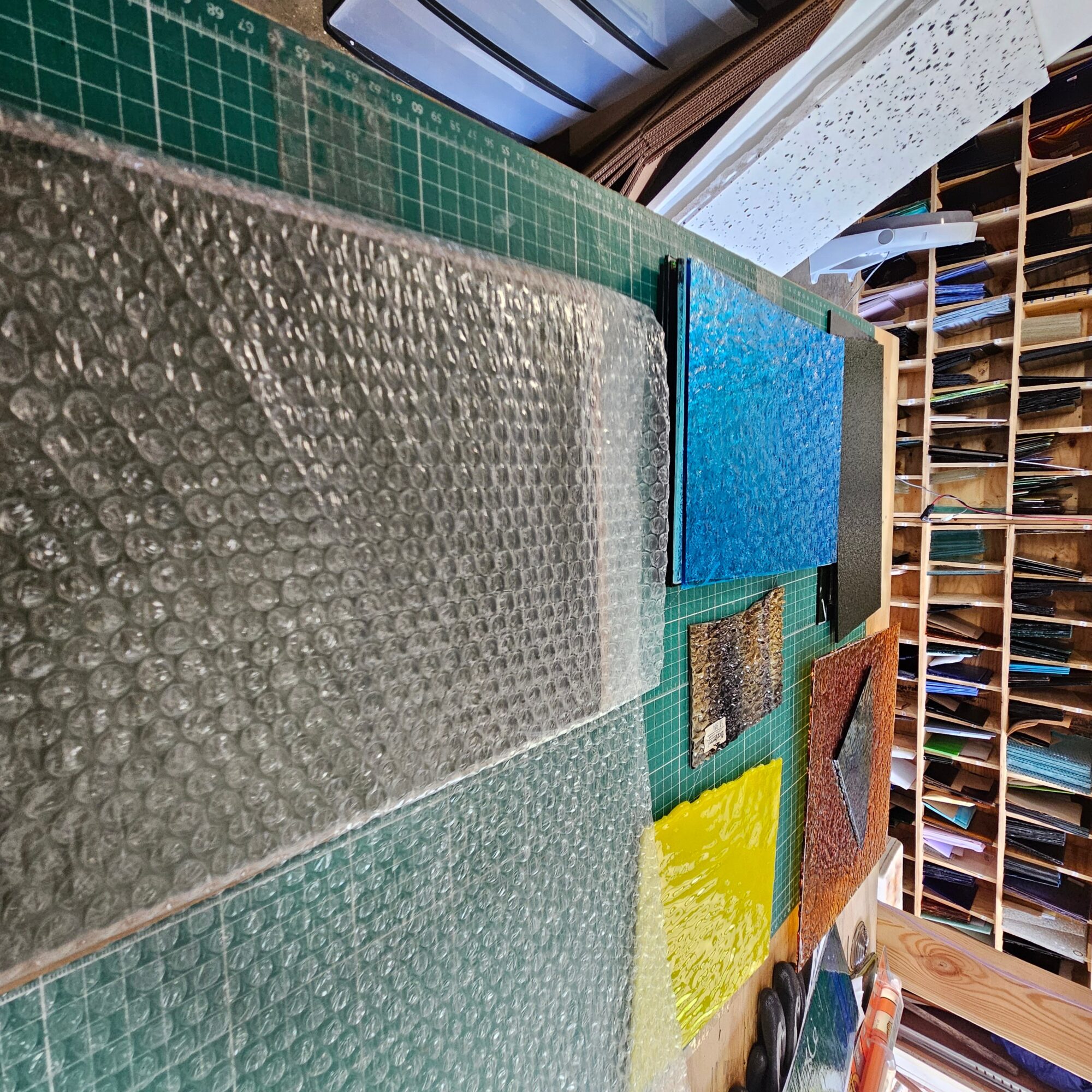 Various textured and colored sheets of material on a green cutting mat, with shelves of paper or fabric in the background.