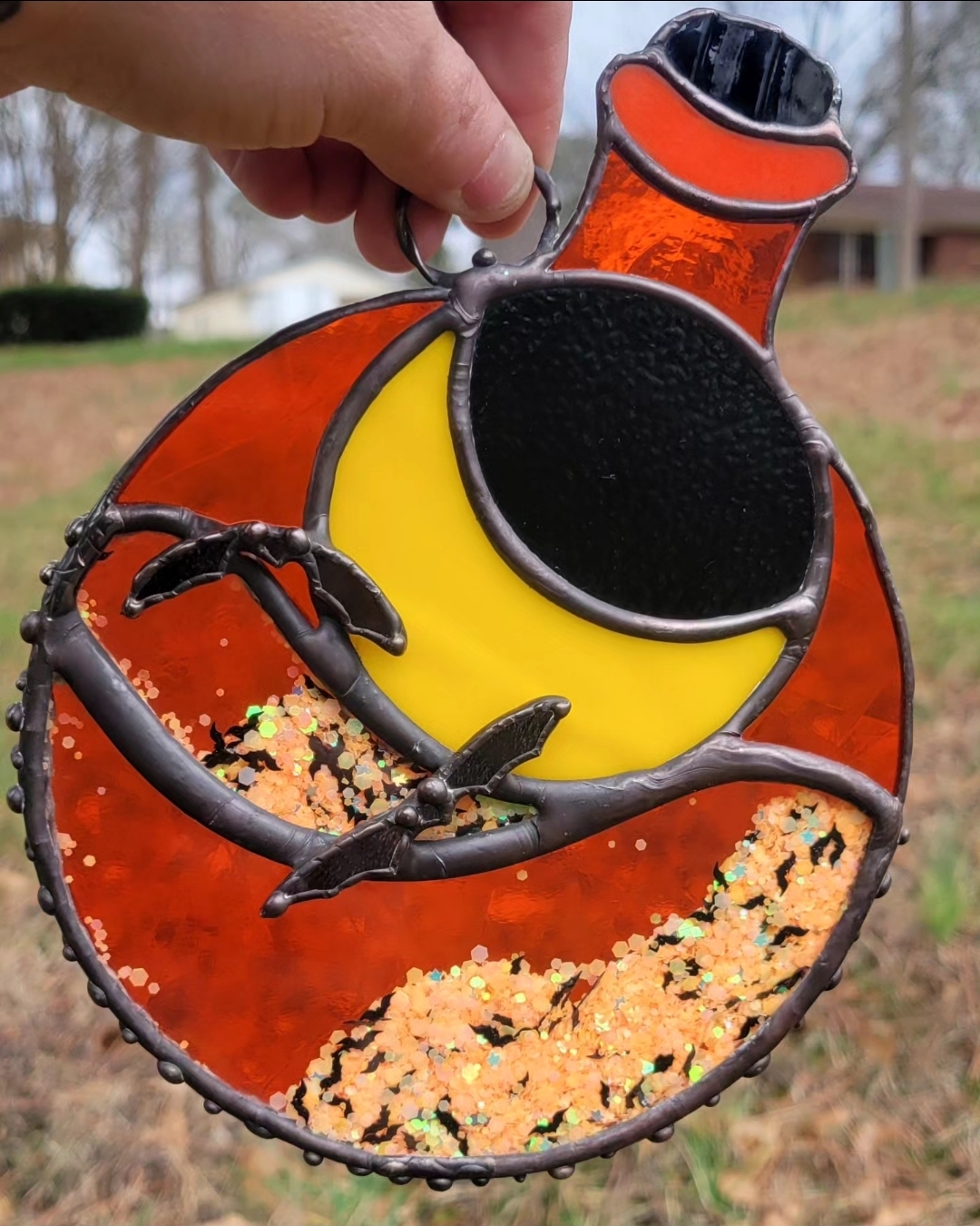 Hand holding a circular stained glass ornament with a red and yellow design, outdoors with trees and a house in background.