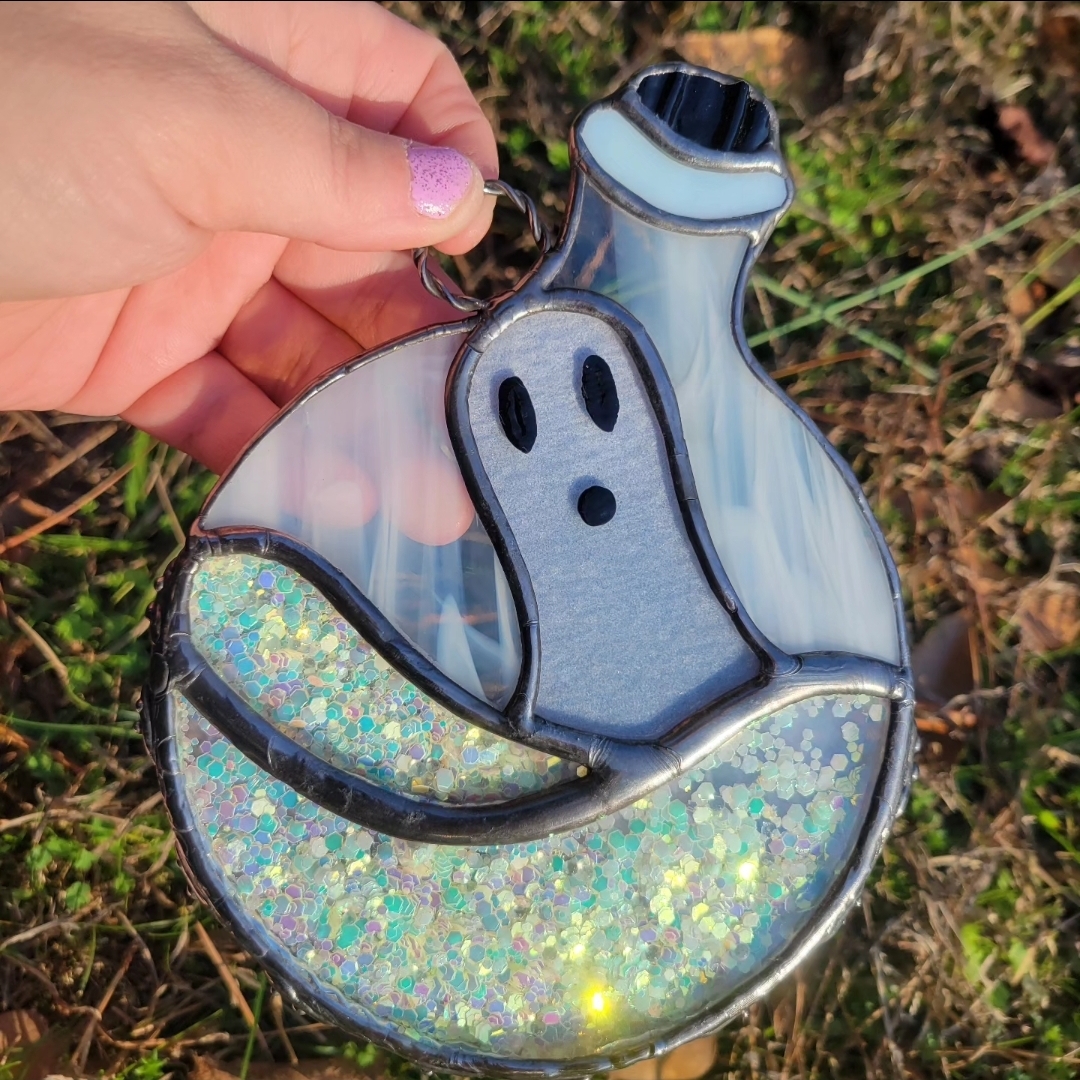 Hand holding a shiny, holographic ghost-shaped object outdoors with grass and leaves in the background.