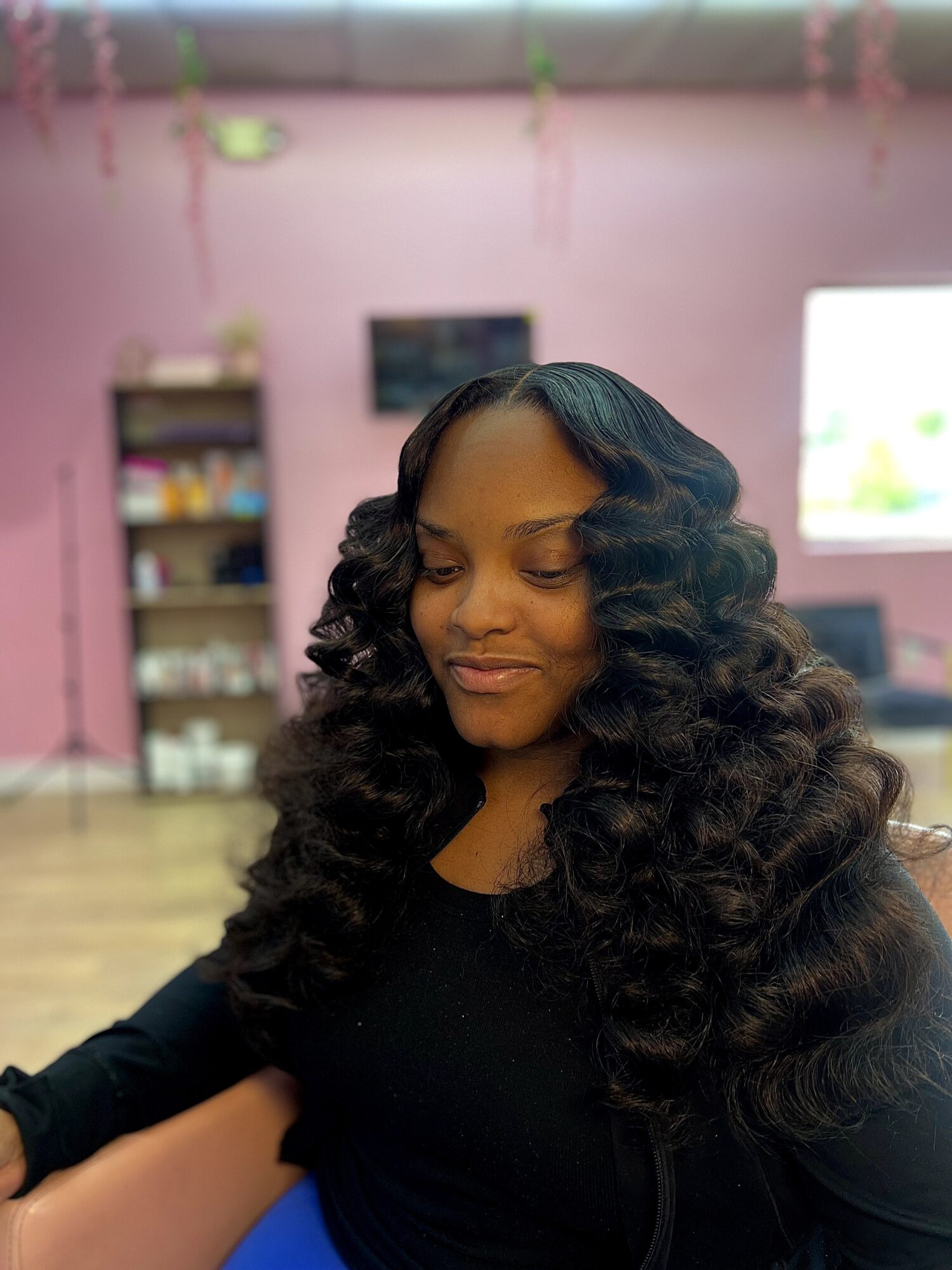 Woman with long, curly hair smiling softly indoors, pink wall and shelves in background.