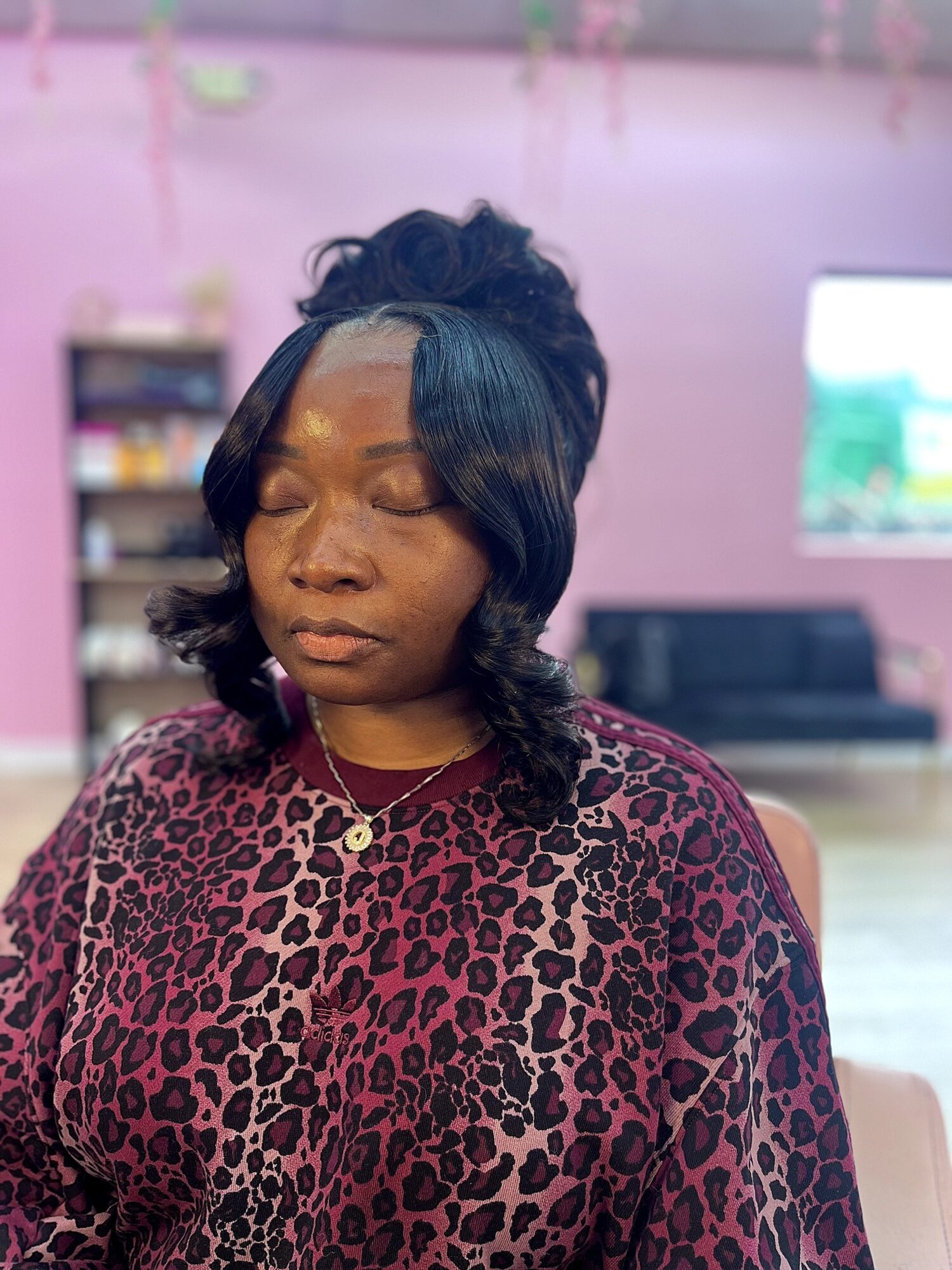 Woman with closed eyes, wearing a leopard print top and a necklace, in a room with pink walls and a TV.