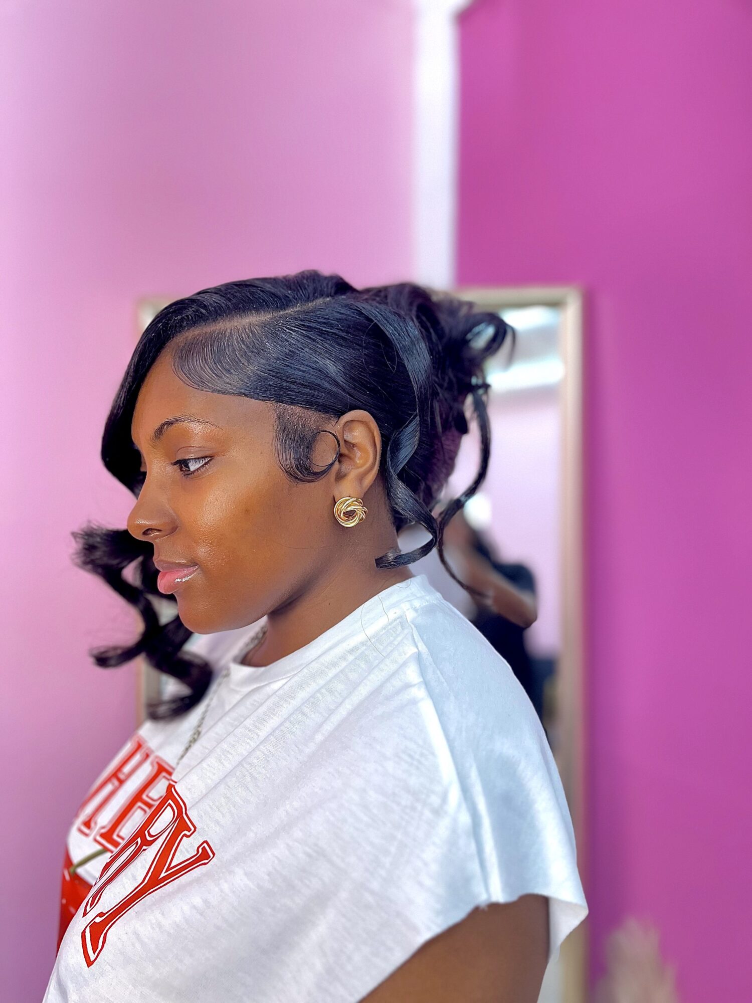 Side profile of a woman with styled hair, earrings, and a white shirt, standing in front of a mirror with a pink wall.