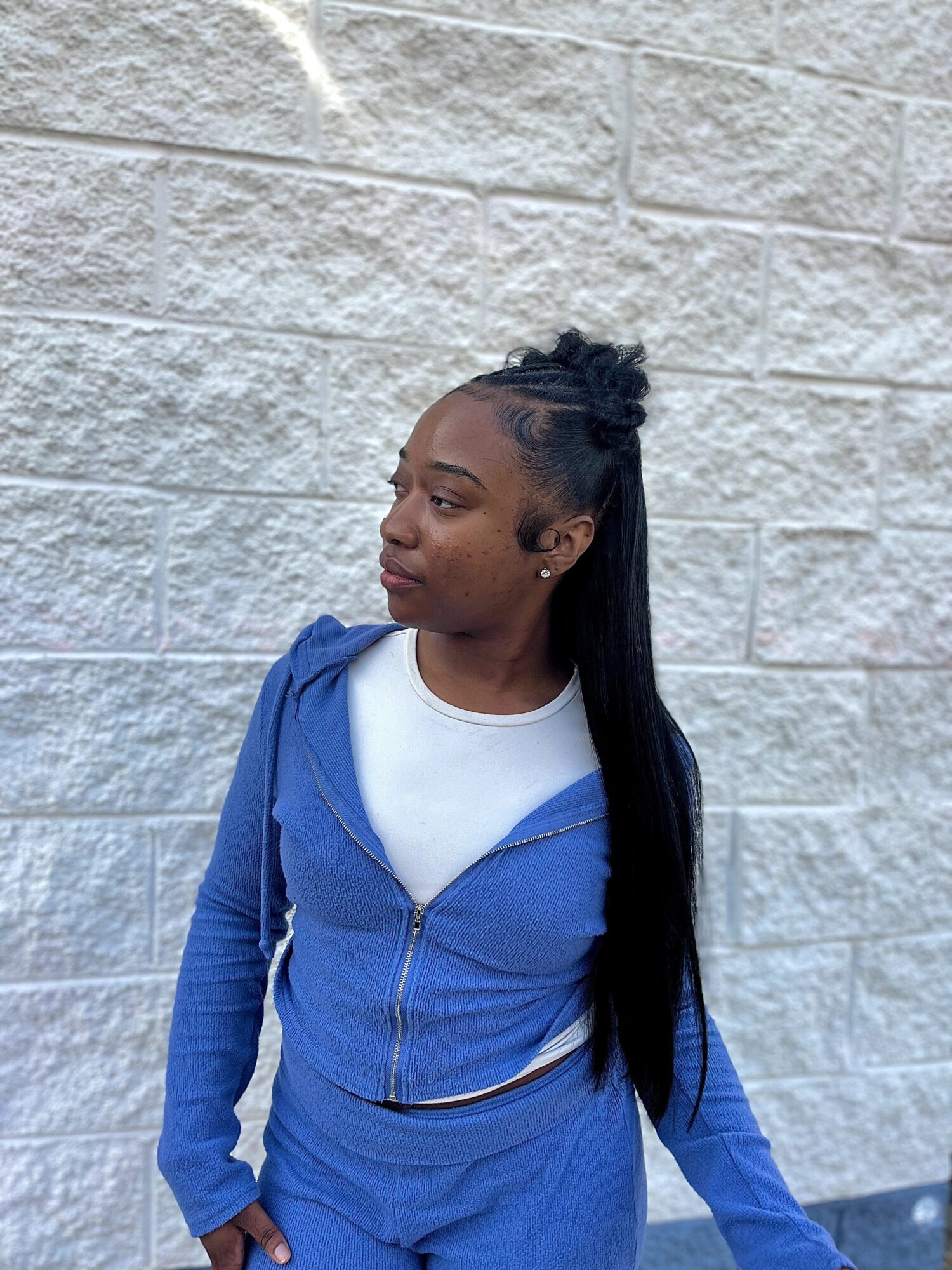 Young woman with long black hair in a high ponytail, wearing a blue zip-up hoodie and white shirt, standing against a brick wall.
