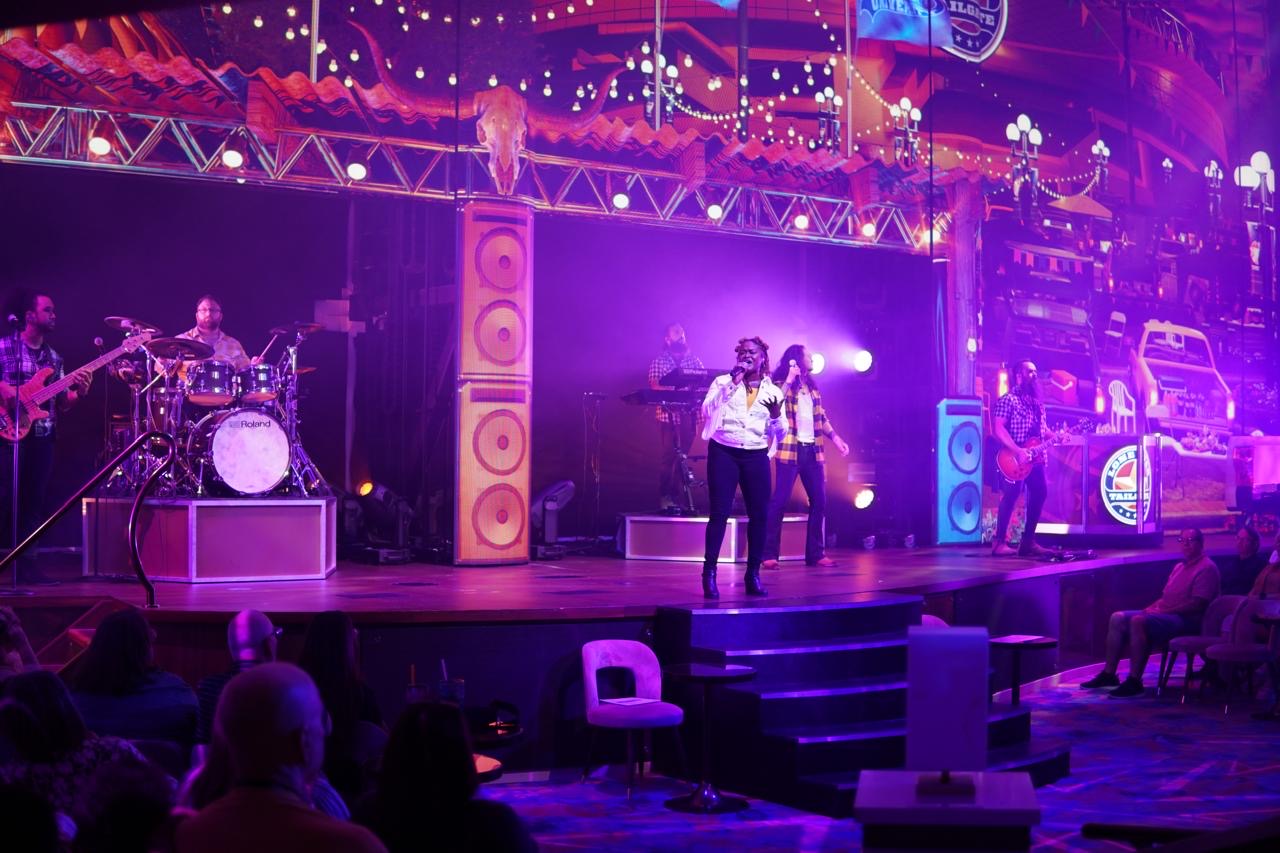 Stage with musicians and singers performing under purple and pink lights, audience watching, colorful speakers, and a multi-level background.