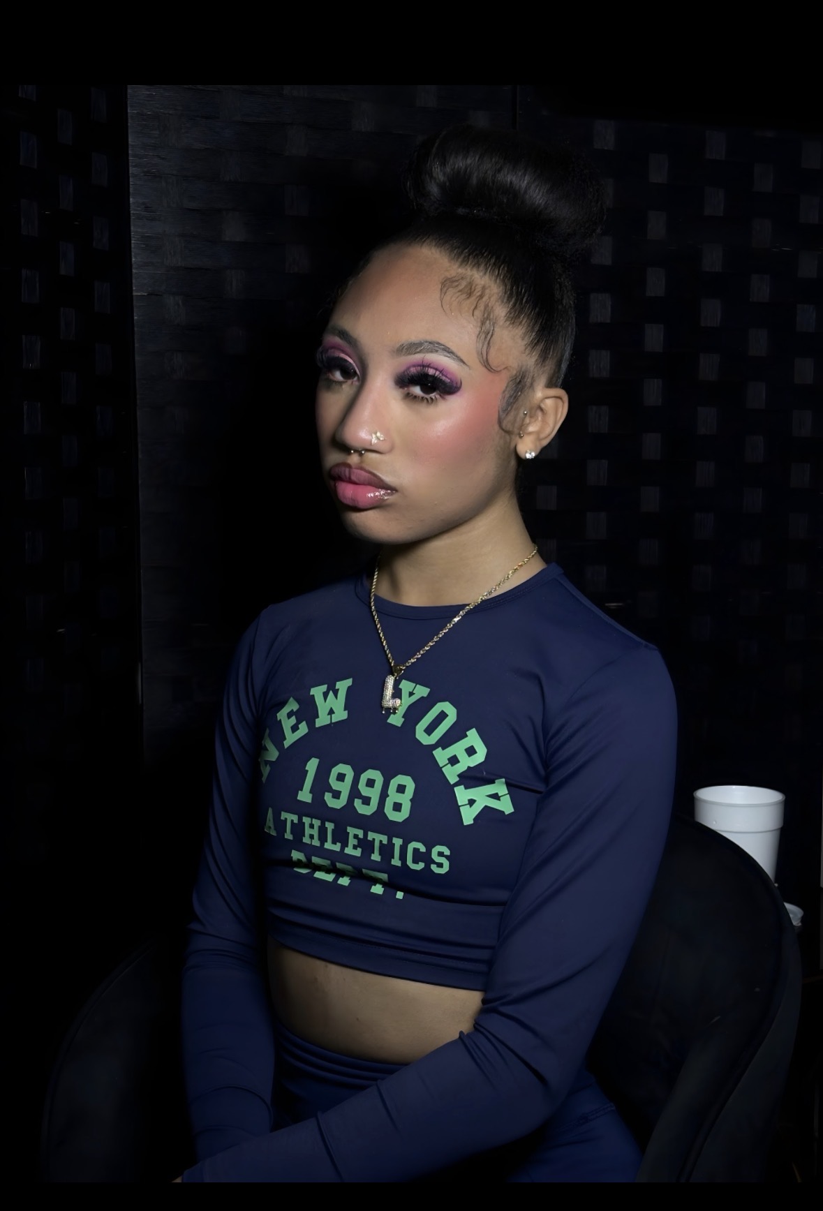 Young woman with styled hair and makeup, wearing a navy crop top with green text, sitting against a dark background.