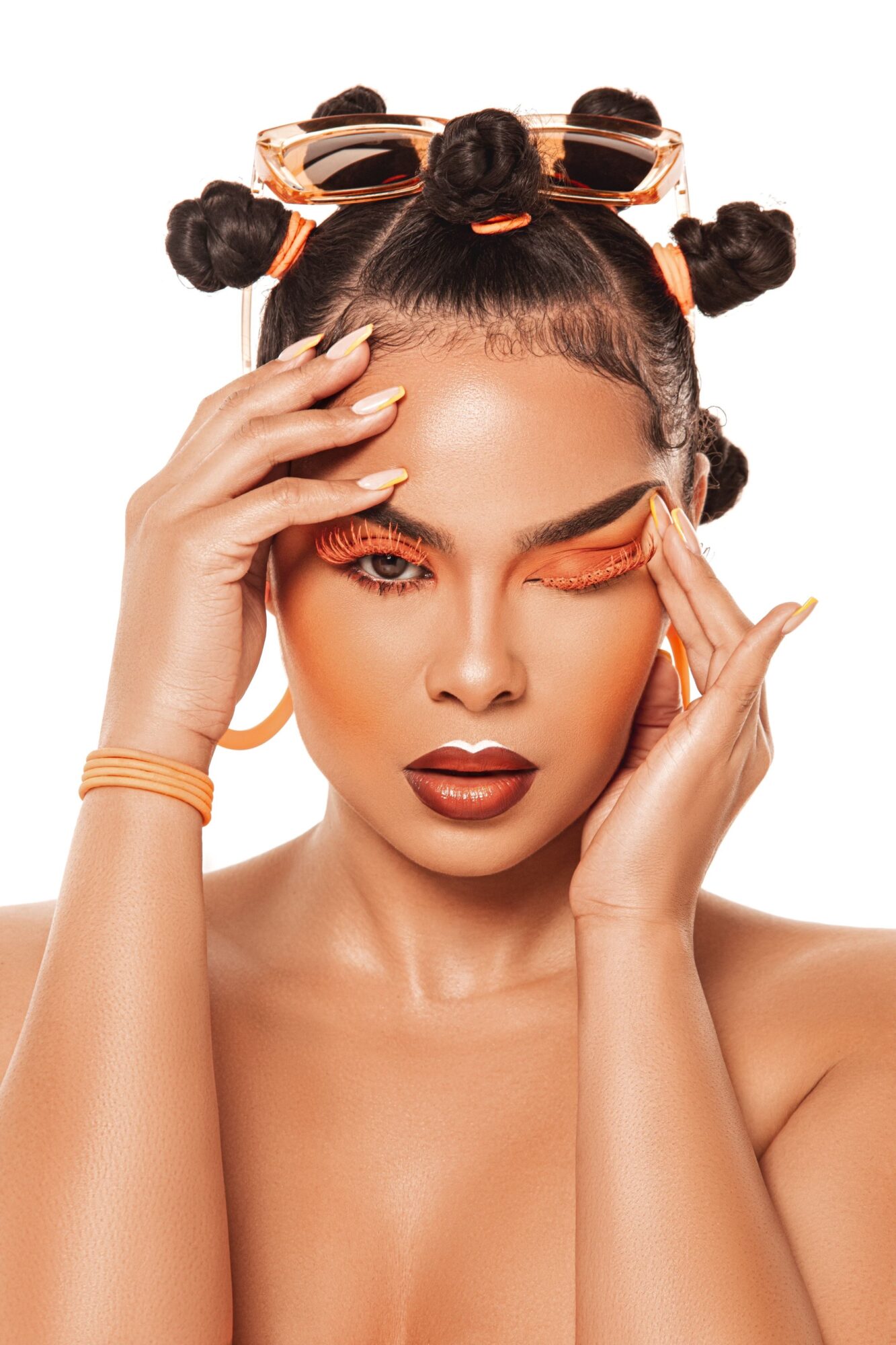 Woman with styled hair, makeup, and sunglasses on head, touching her face with both hands, looking at the camera.