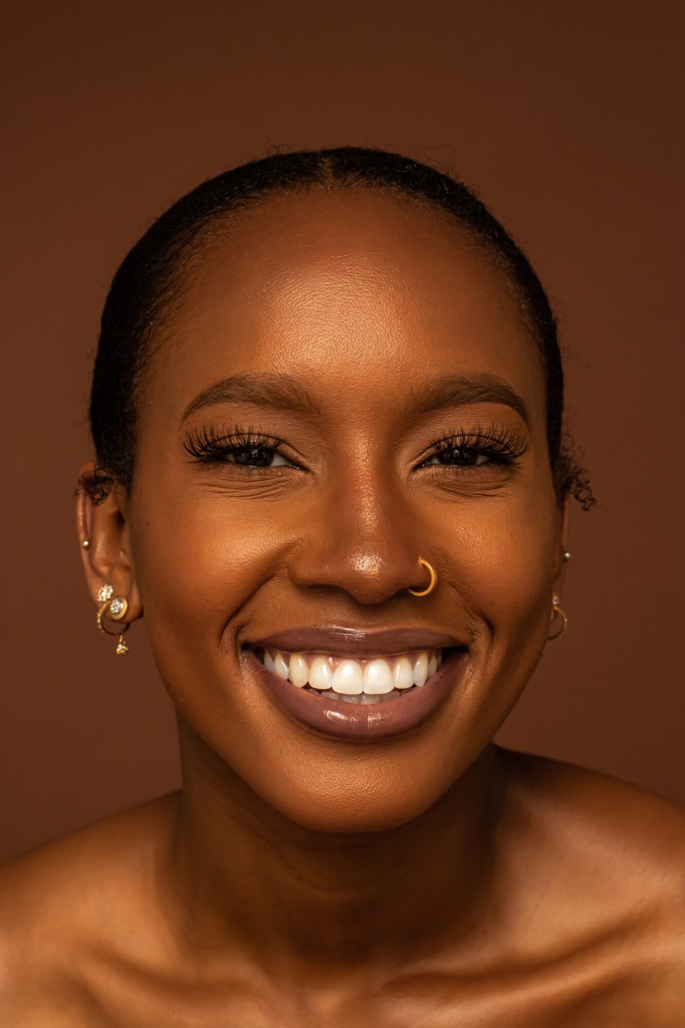 Close-up of smiling woman with dark skin, wearing earrings and a nose ring, against a brown background.