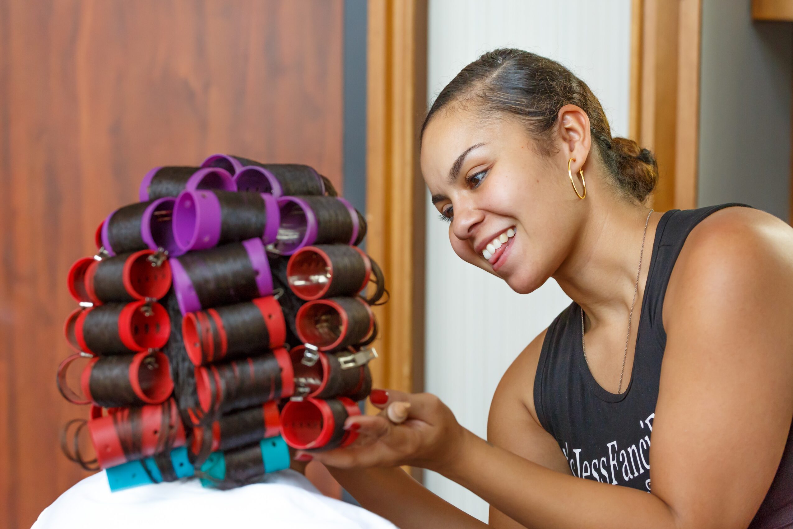 Smiling woman with curly hair in a black tank top looks at colorful hair rollers stacked on a wall, indoors.