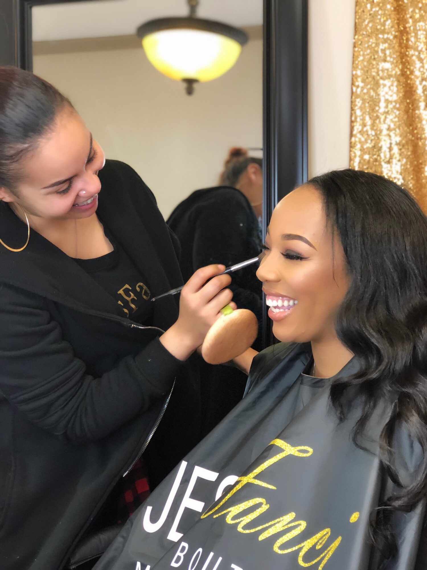 Makeup artist applying makeup to smiling woman with long dark hair, in a room with gold curtains and a mirror.