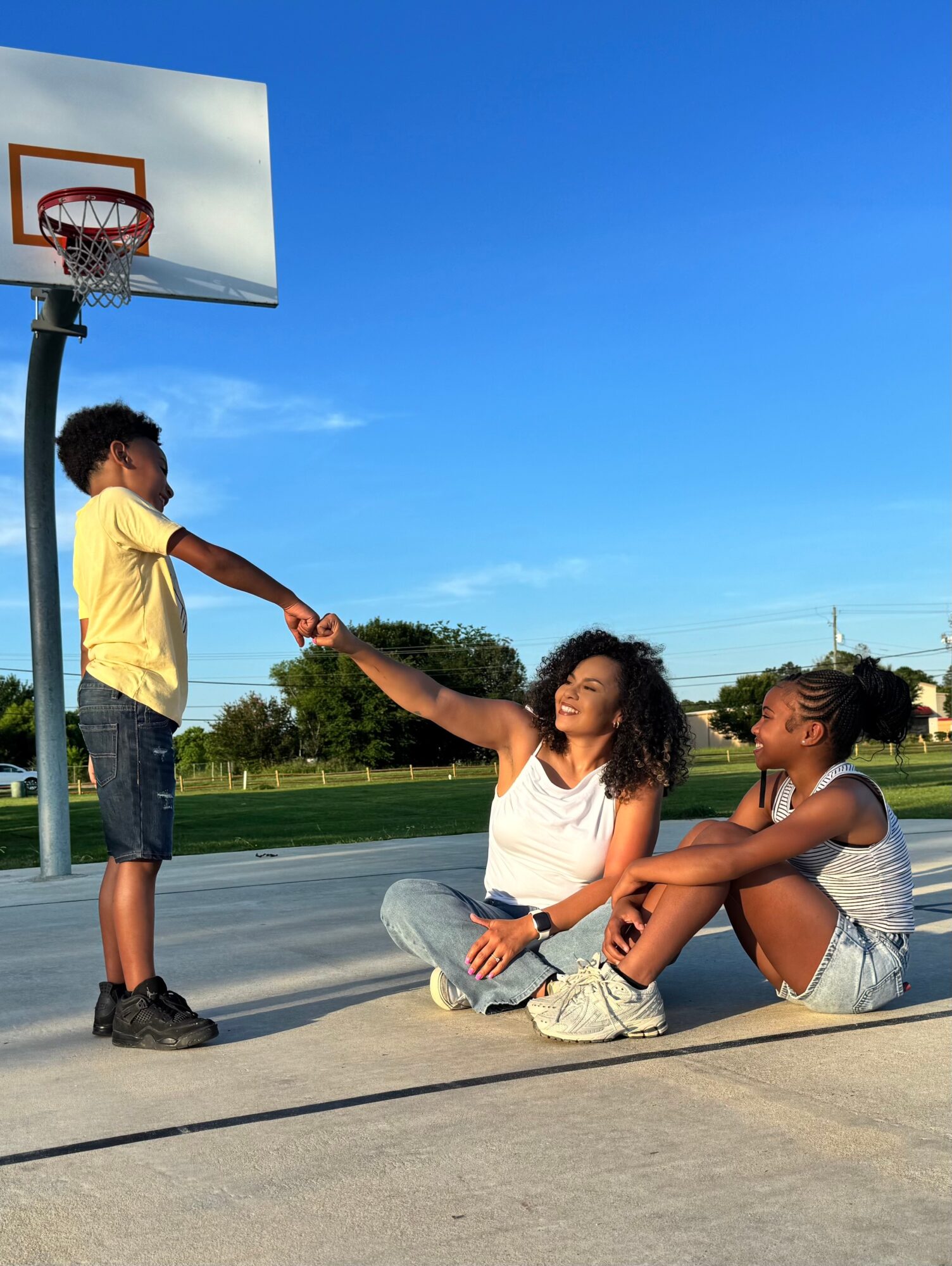 Two girls sitting on the ground and a boy standing near a basketball hoop outdoors on a sunny day.