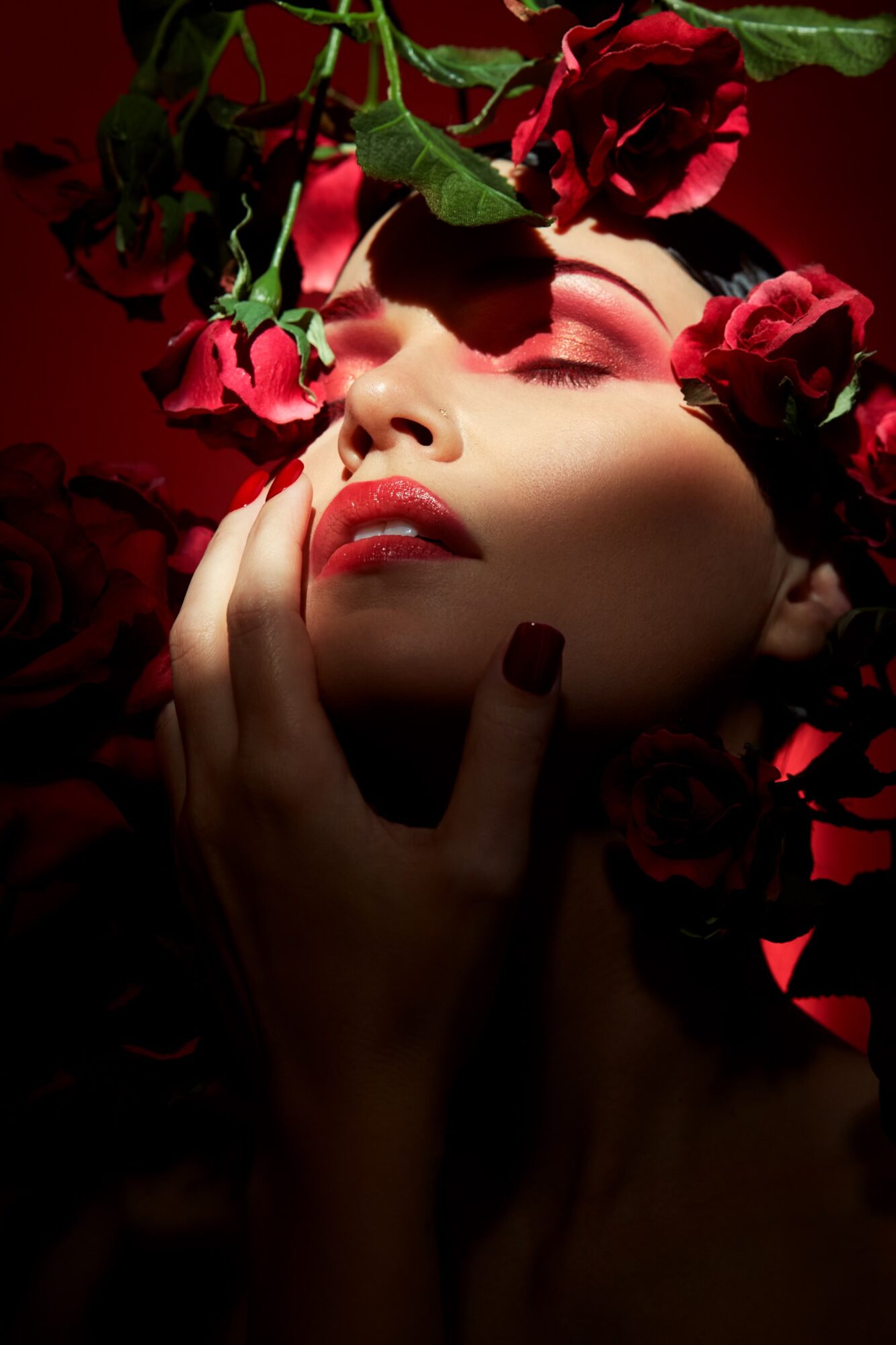 Close-up of a woman with eyes closed, surrounded by red roses, with hand touching her face, dark background.