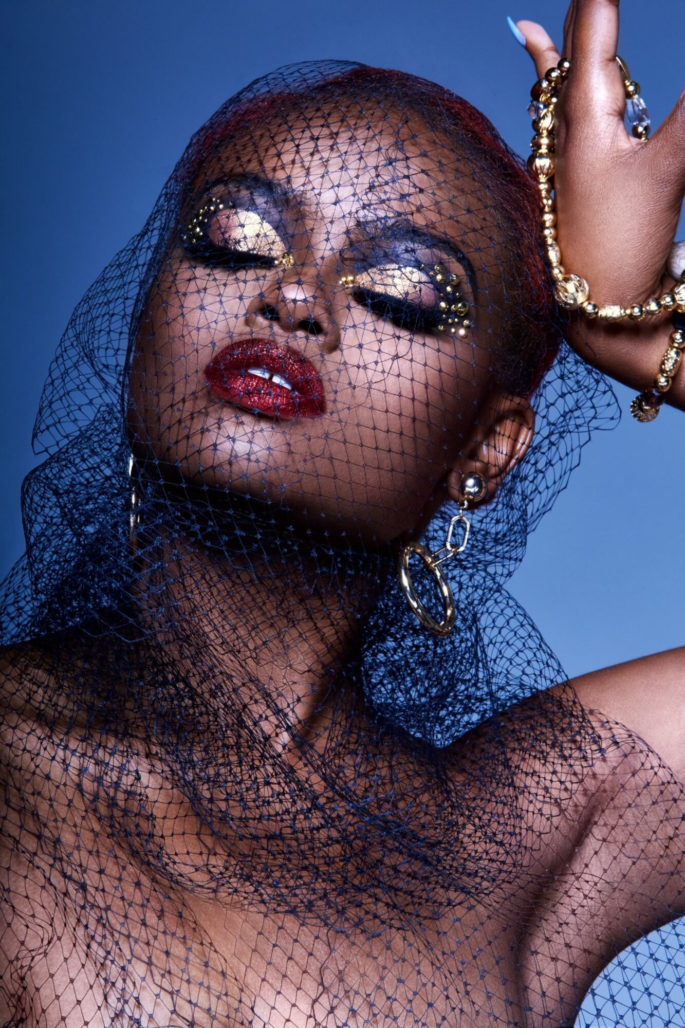 Close-up of a woman with glittery makeup, wearing jewelry and a black net veil, against a blue background.