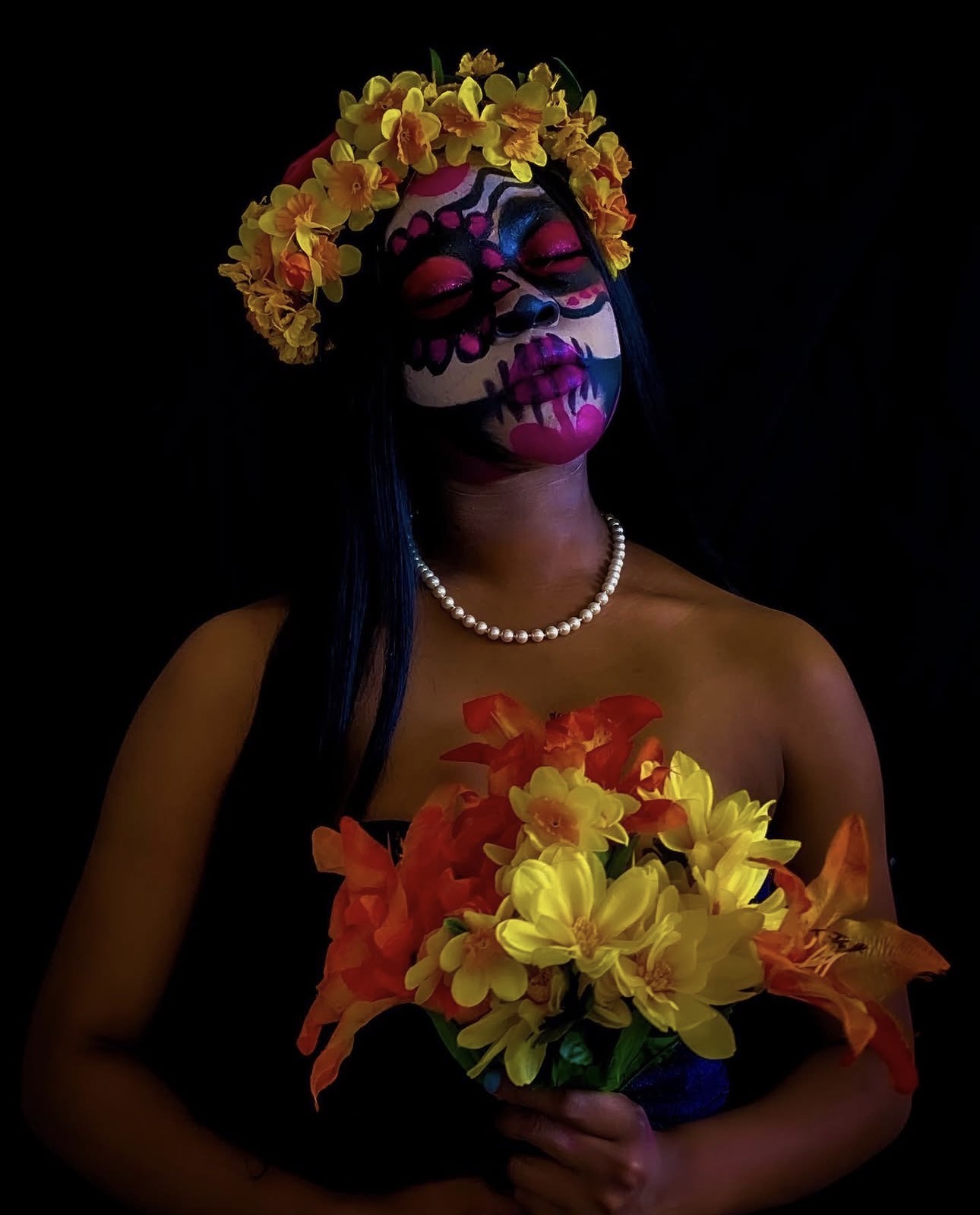 Person with sugar skull face paint, wearing a yellow flower crown, holding a colorful bouquet, against a black background.