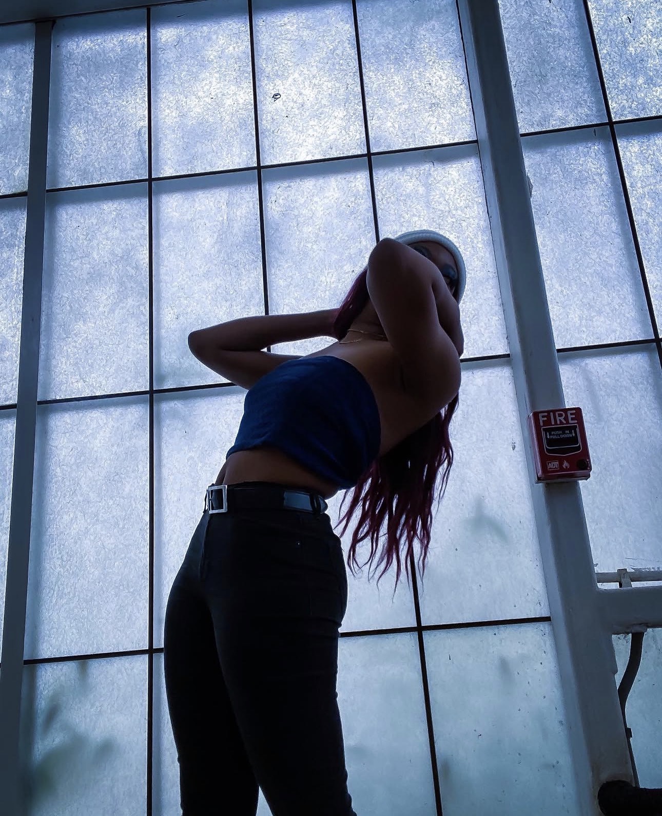 Person standing with arms raised, back to camera, in front of large frosted glass window, with fire alarm on wall.