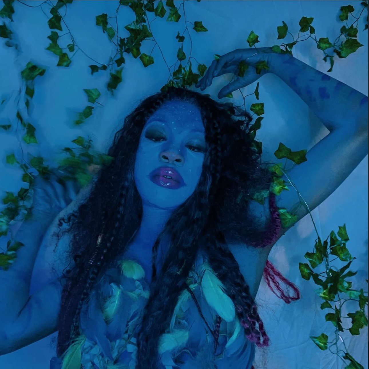 Woman with curly hair lying among green vines, illuminated by blue light, with one arm raised above her head.