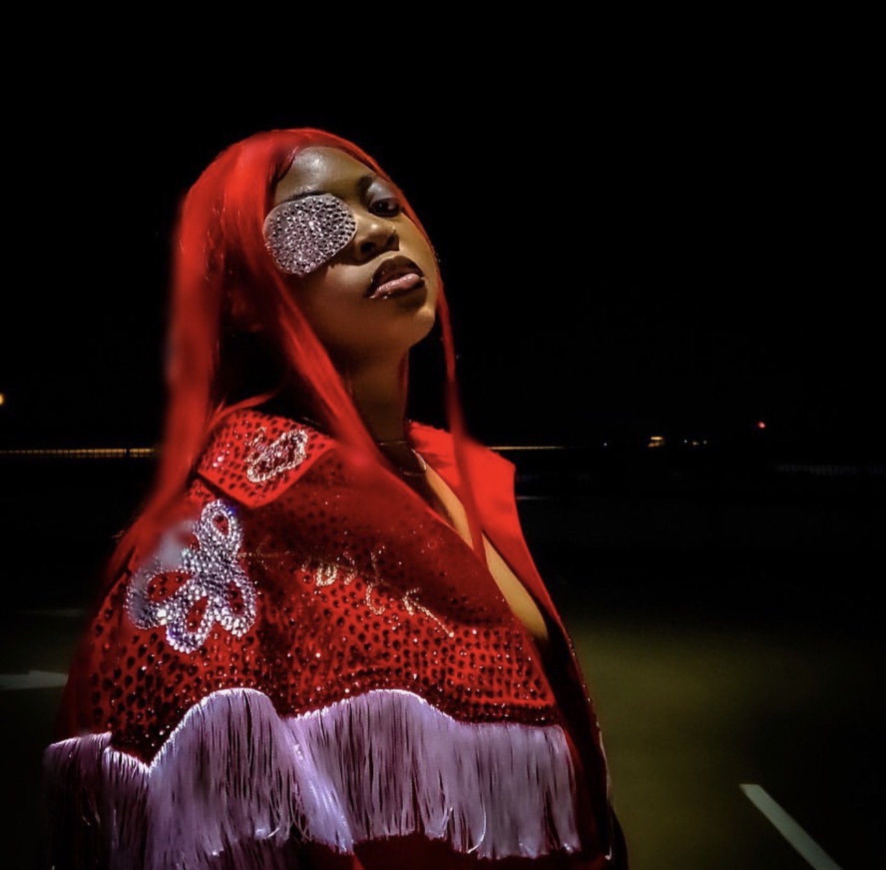 Person with long red hair wearing a red and purple sequined outfit and an eye patch, standing outdoors at night.
