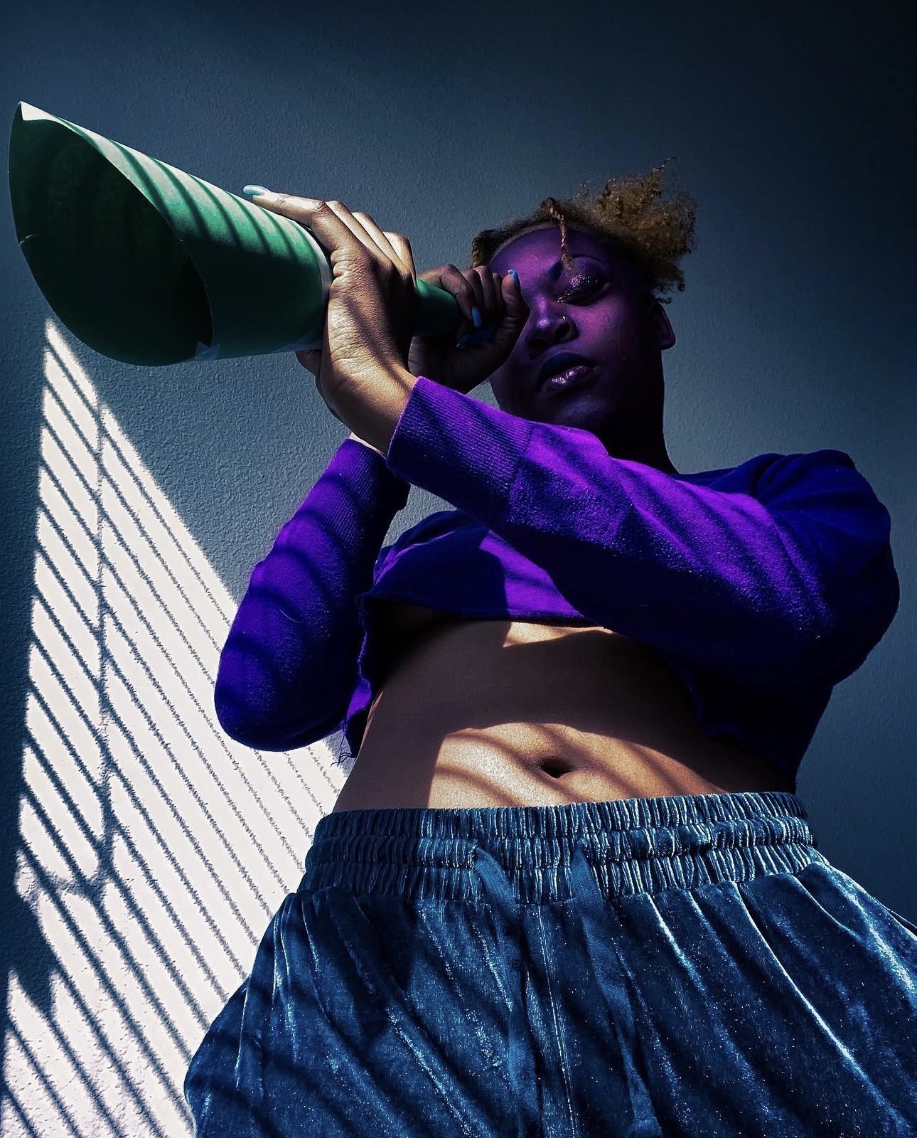 Person with purple clothing holding a green object, shadow cast by blinds on wall, dark background, low angle shot.