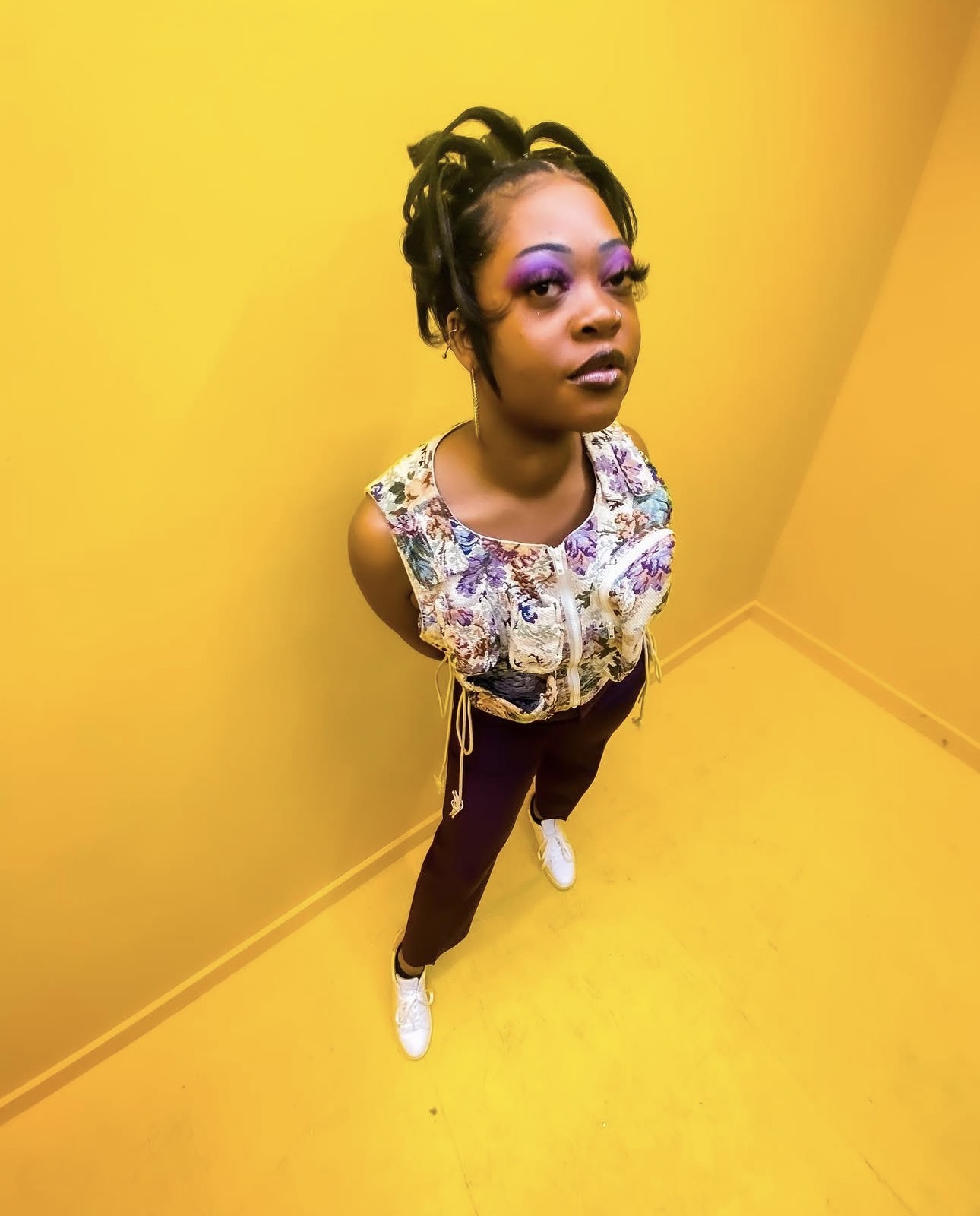 Young woman with styled hair and colorful makeup stands against a yellow wall, looking at the camera with a serious expression.