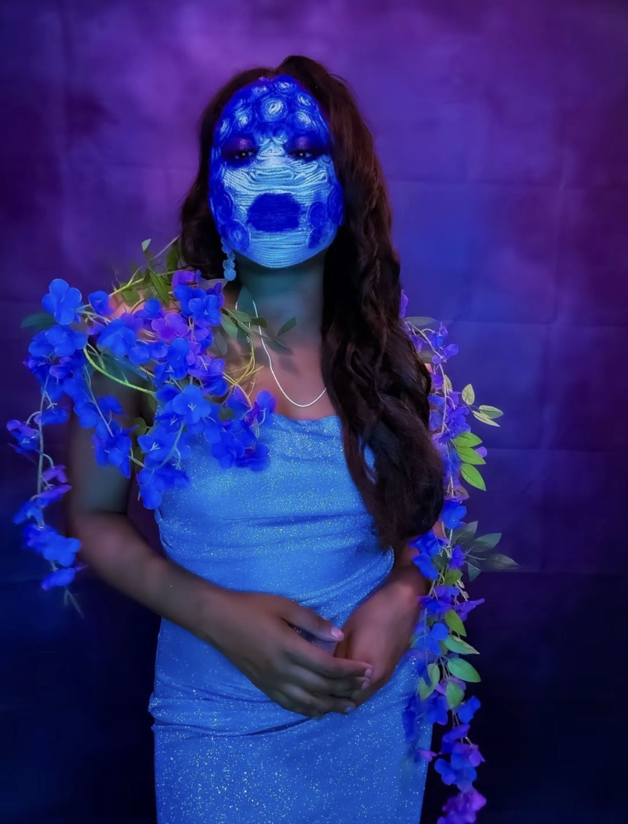 Person wearing a blue dress and a mask with a blue face design, holding blue flowers, against a purple background.