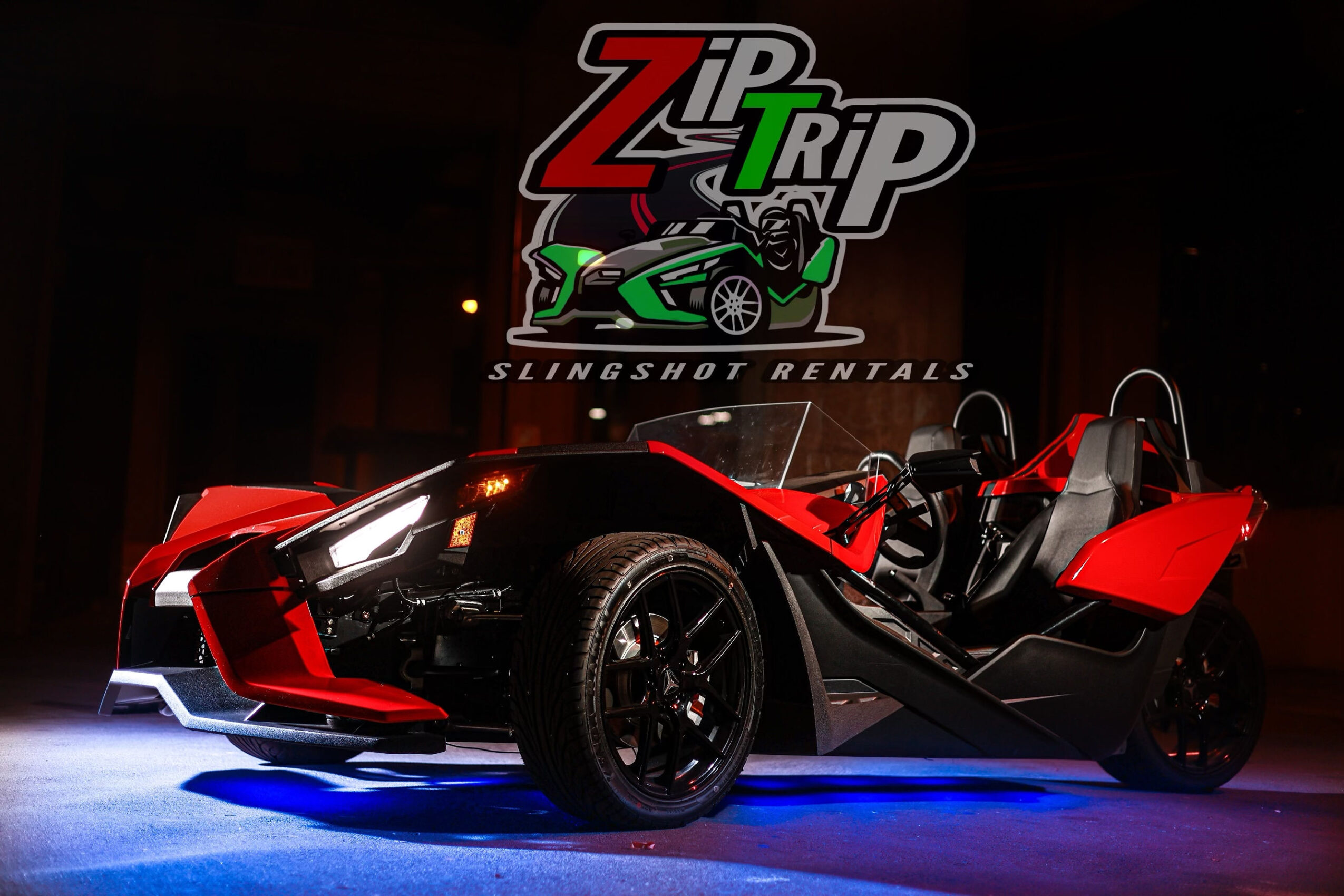 Red and black three-wheeled vehicle with a canopy, displayed indoors with a Zip Trip Slingshot Rentals sign above.