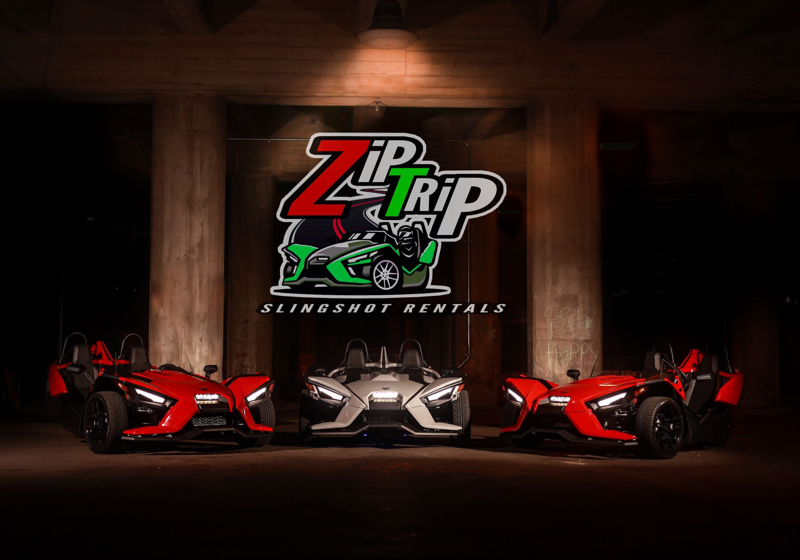 Three cars are displayed in a dimly lit indoor setting with a sign reading 'Zip Trip Slingshot Rentals' behind them.
