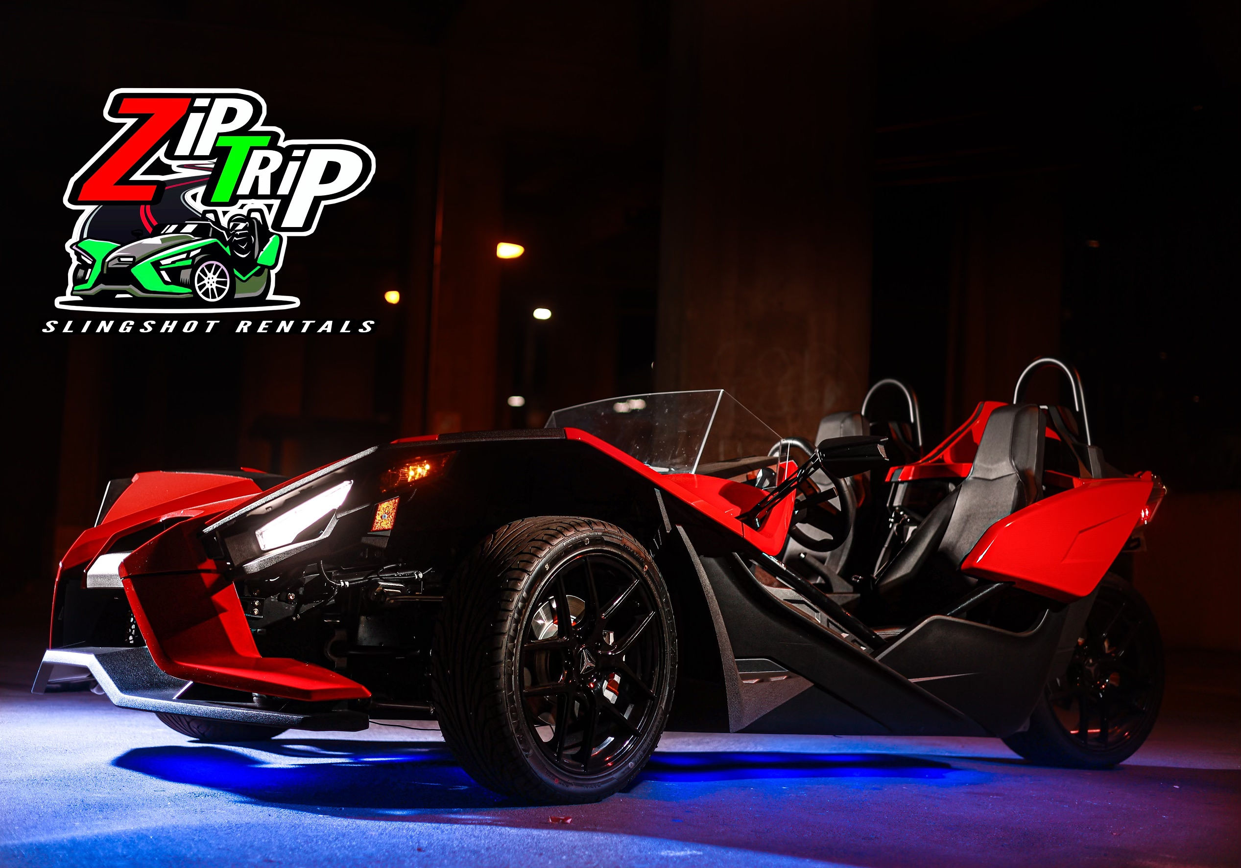 Red and black three-wheeled vehicle on stage with Zipp Slingshot Rentals logo above, dark background, illuminated by stage lights.
