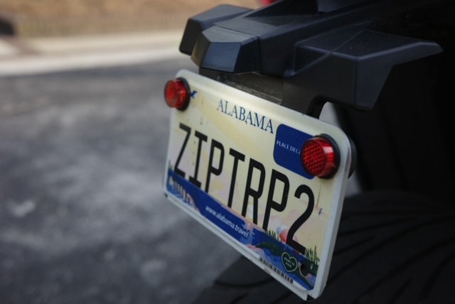 Alabama license plate with red lights on a vehicle, reading ZIPTRO2, mounted on the rear of a motorcycle or vehicle, with a blurred background.