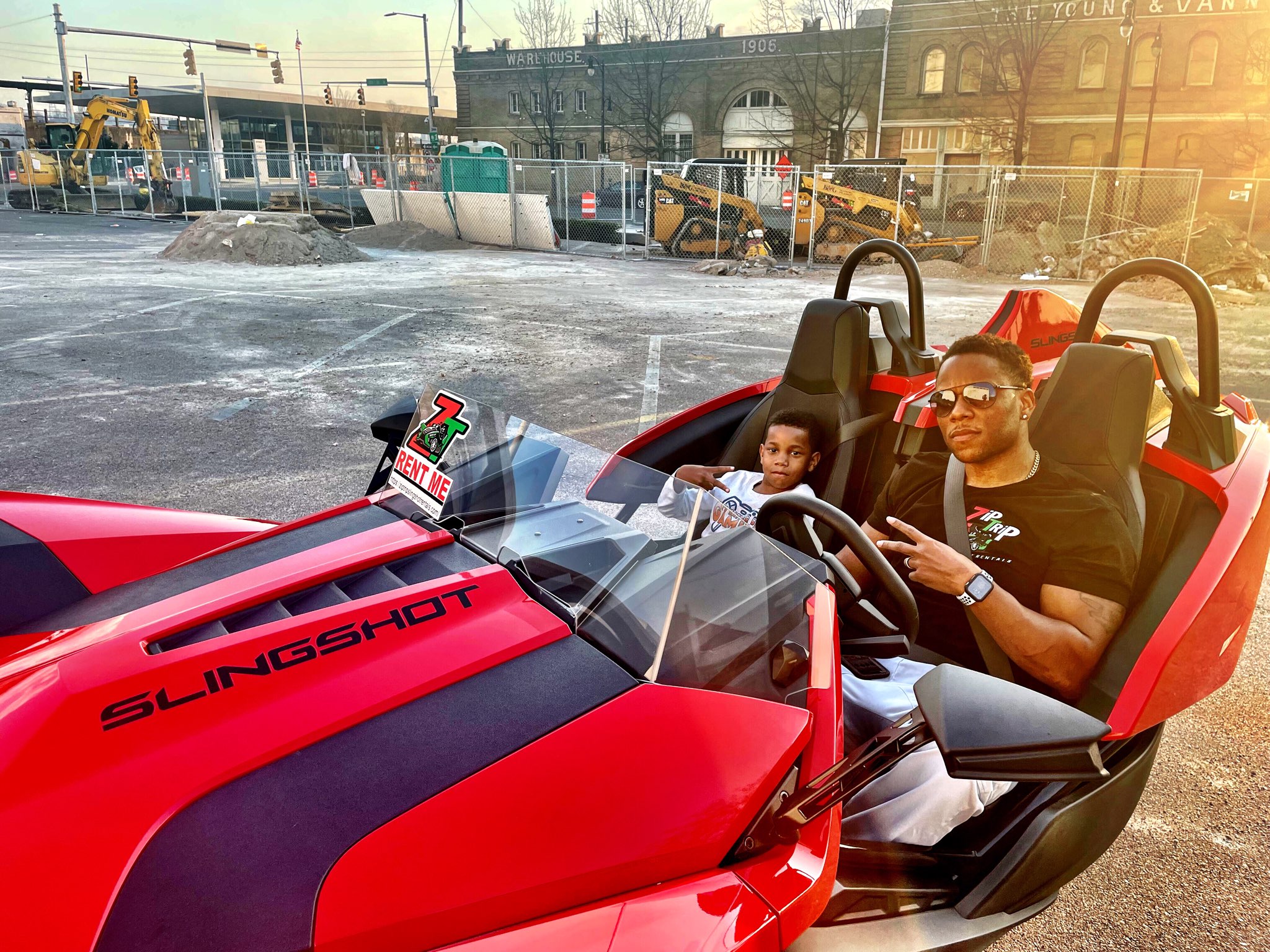Person driving a red and black three-wheeled vehicle with a child passenger, in an outdoor parking lot.